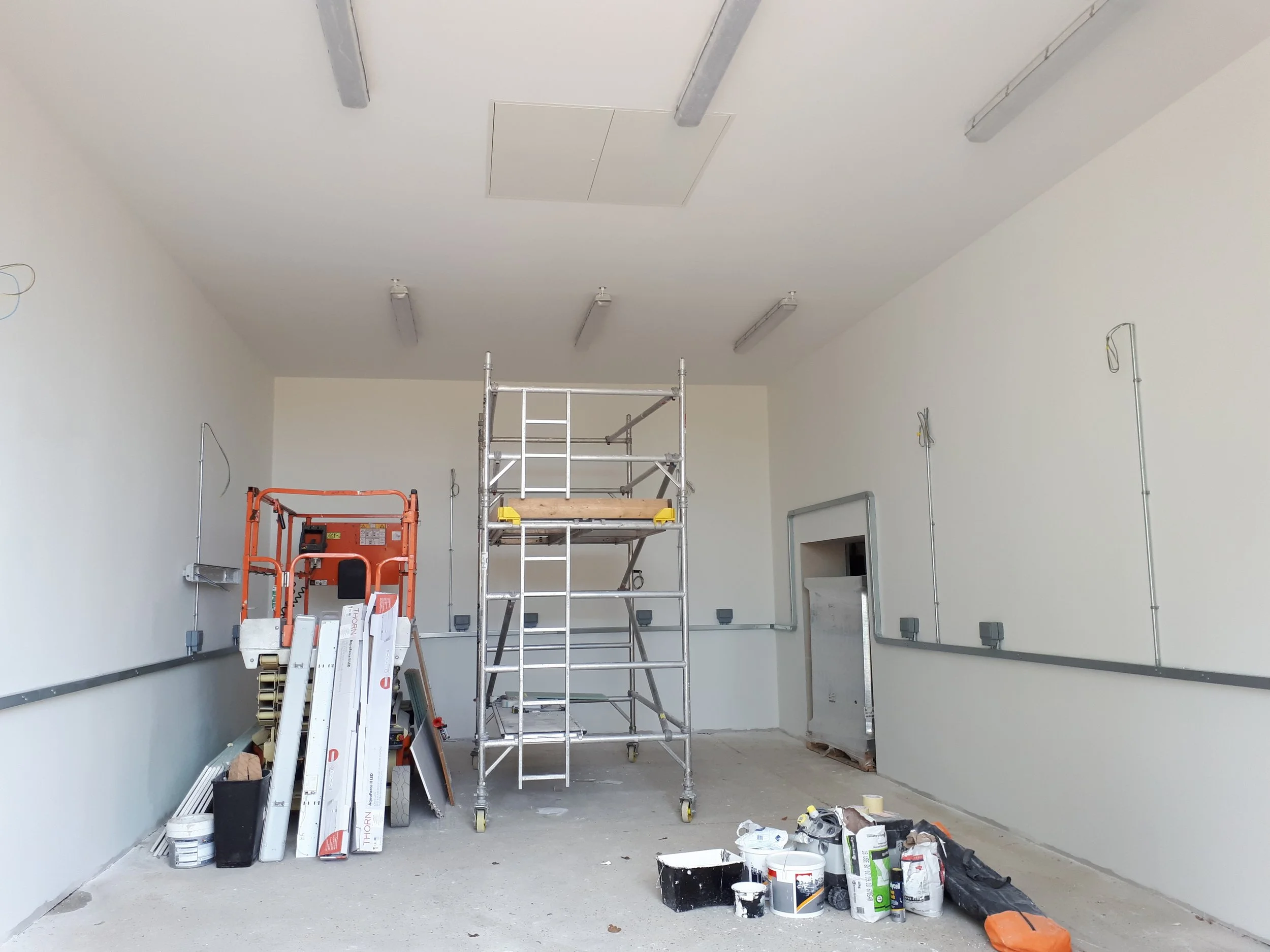Interior of a room under renovation with scaffolding, ladders, paint buckets, and construction supplies scattered on the floor and against the walls, which are painted white.