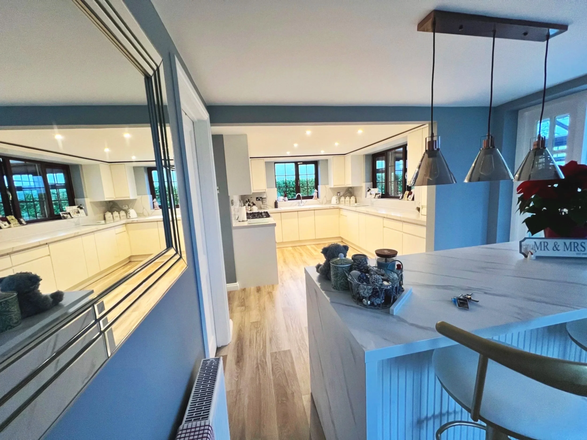 View of a modern kitchen with white cabinetry, large windows, and pendant lights. Part of a white island with decorative items and a poinsettia plant is visible in the foreground. The room has hardwood flooring and recessed ceiling lights.
