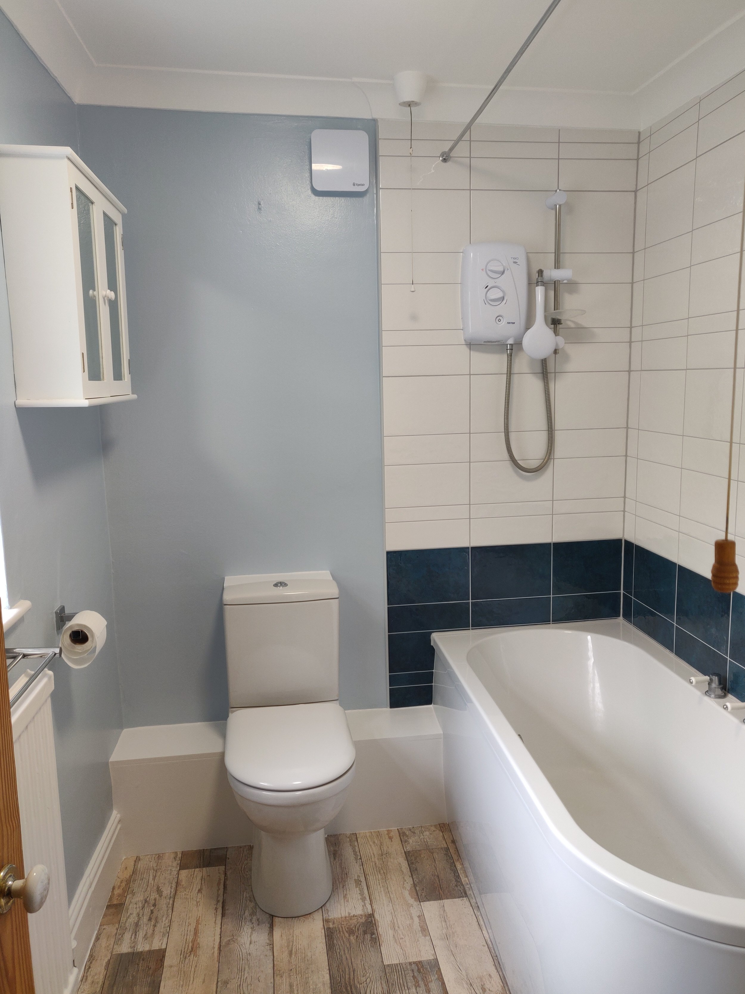 A small bathroom with a white toilet, a bathtub with blue and white tiled walls, a wall-mounted electric water heater, a showerhead, a white cabinet, a toilet paper holder, and wooden flooring.