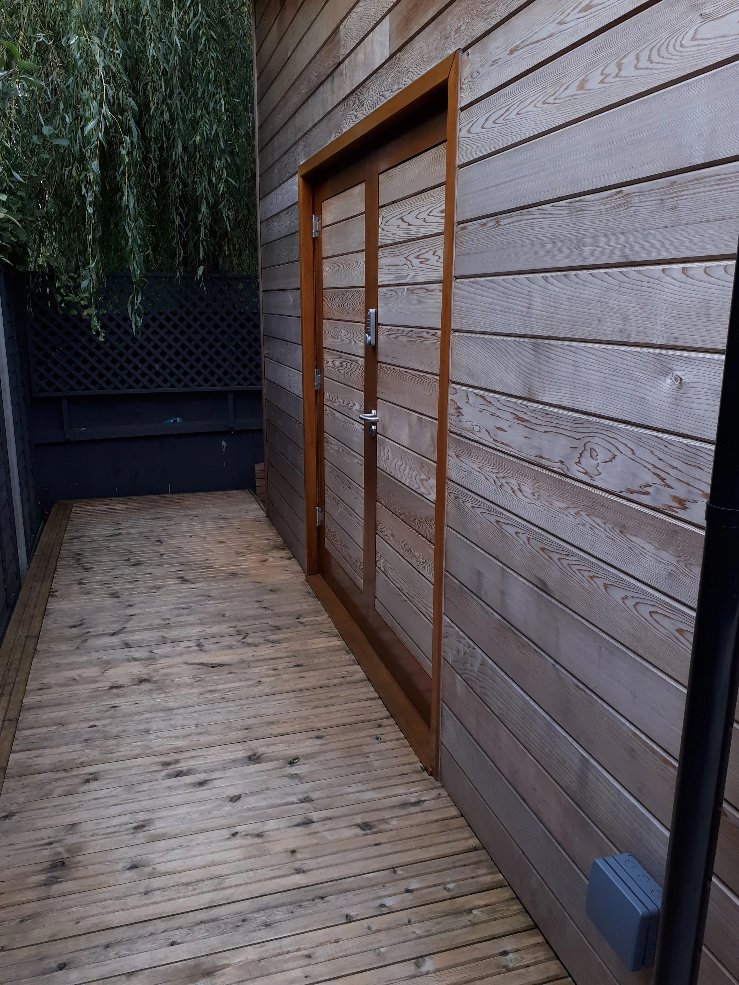 Wooden outdoor deck with a wall of horizontal wooden planks and a small door, surrounded by greenery and a black lattice fence.