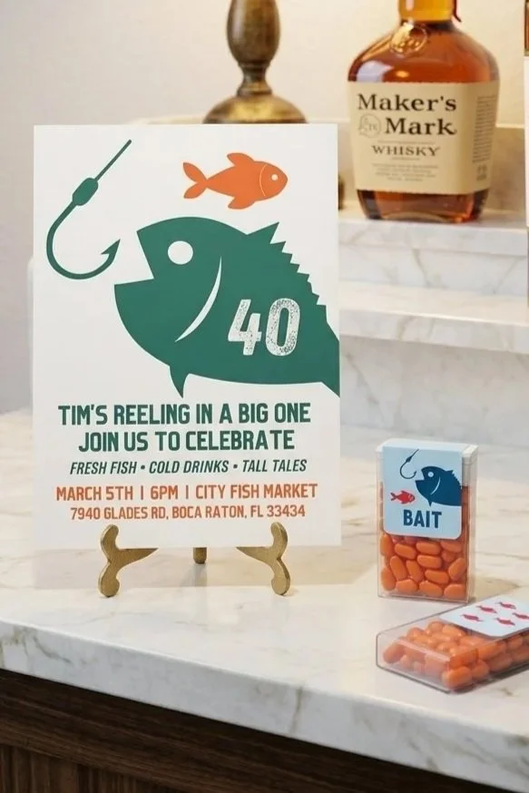 A fishing-themed 40th birthday invitation sign featuring a fish graphic with the number 40, a fish bait container, and a bottle of Maker's Mark whisky on a marble table.