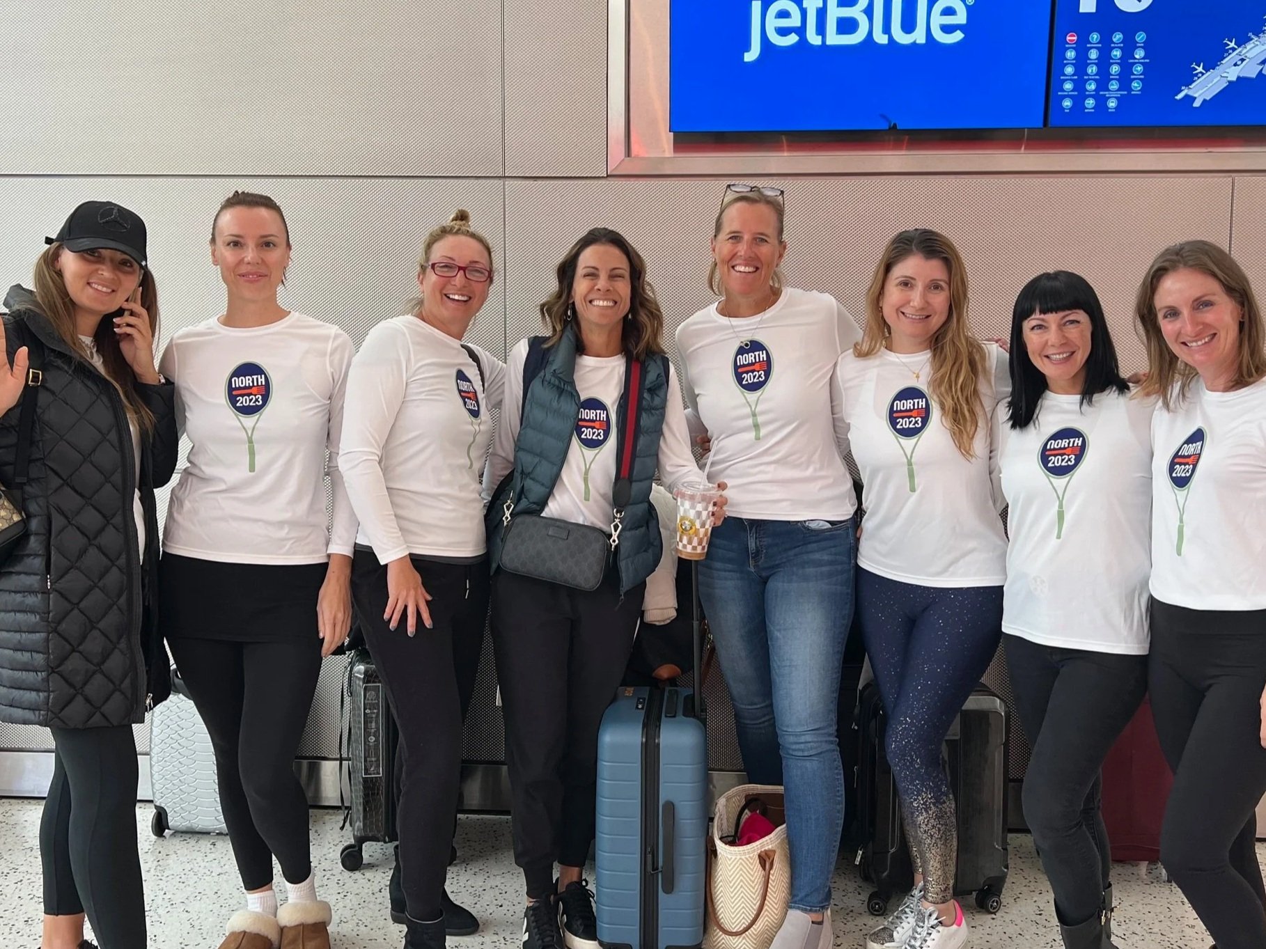 A group of eight women smiling at an airport, standing in front of luggage, with a blue JetBlue sign overhead. They are wearing white shirts with 'North 2023' printed on them, and some are holding or surrounded by travel bags.