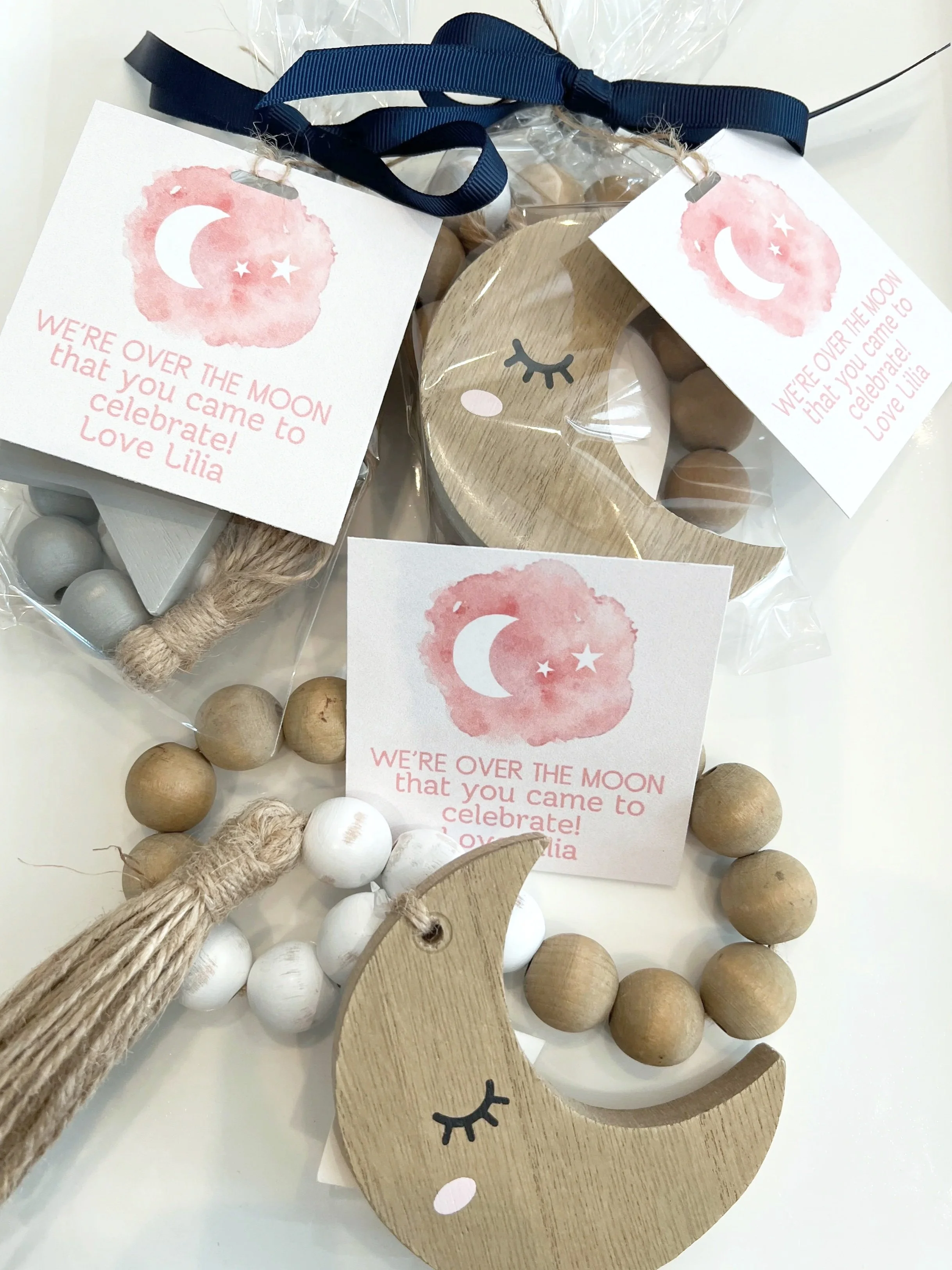 A decorative wooden crescent moon with closed eyes and long eyelashes, surrounded by wooden and white beads, and two printed tags with pink watercolor circles, crescent moon and stars, and text reading 'We're over the moon that you came to celebrate!