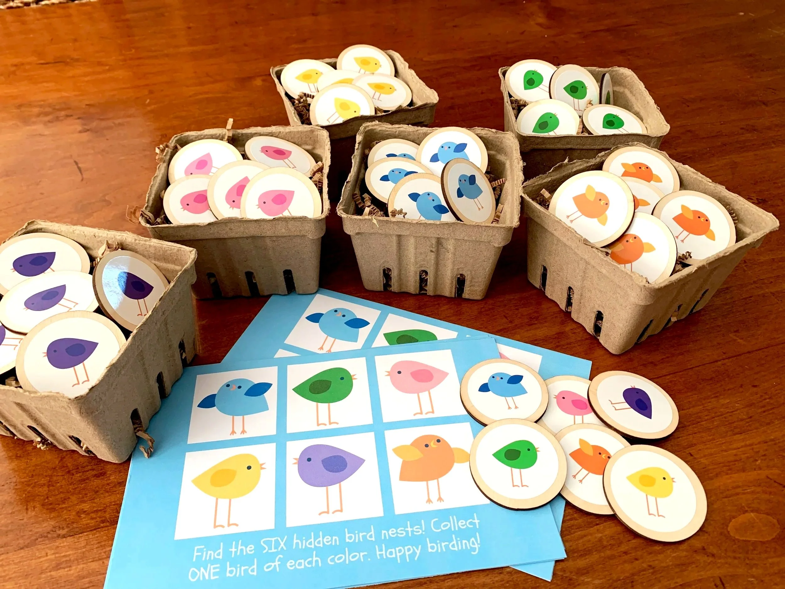 Several containers of stickers depicting colorful birds, including pink, blue, green, purple, yellow, and orange, are displayed on a wooden table along with a blue card with instructions for a bird-themed activity.