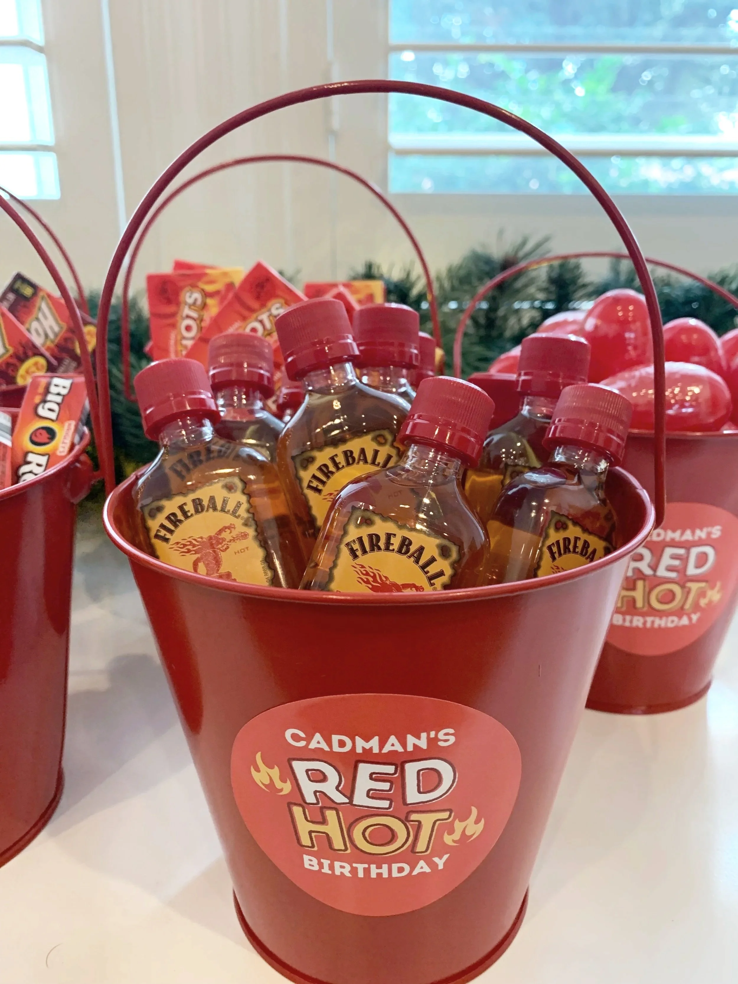 Red bucket containing Fireball hot cinnamon whiskey miniature bottles with a Cadman's Red Hot birthday sticker, seen on a white table near a window.