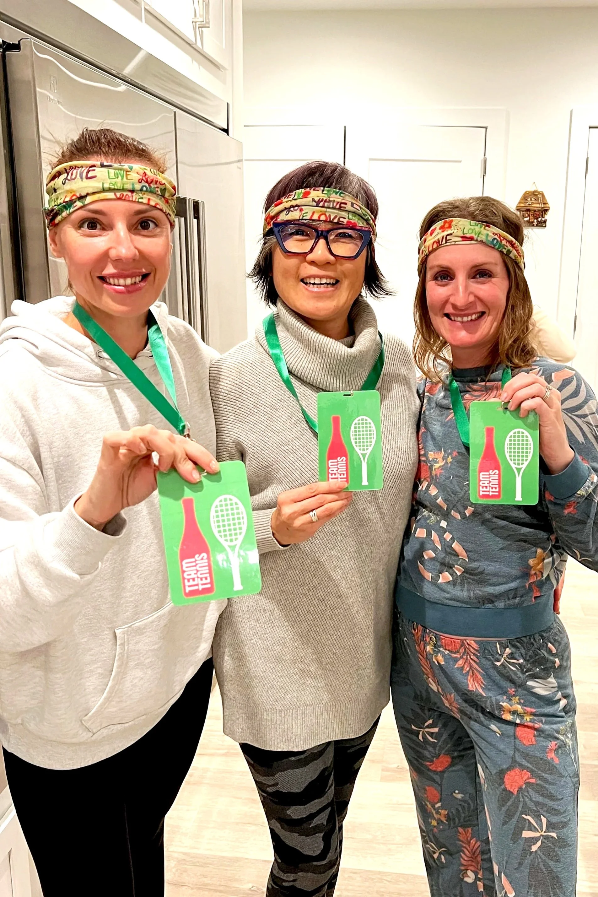 Three women wearing colorful headbands with the word 'LOVE' and holding green medals with tennis racket icons, indoor setting.