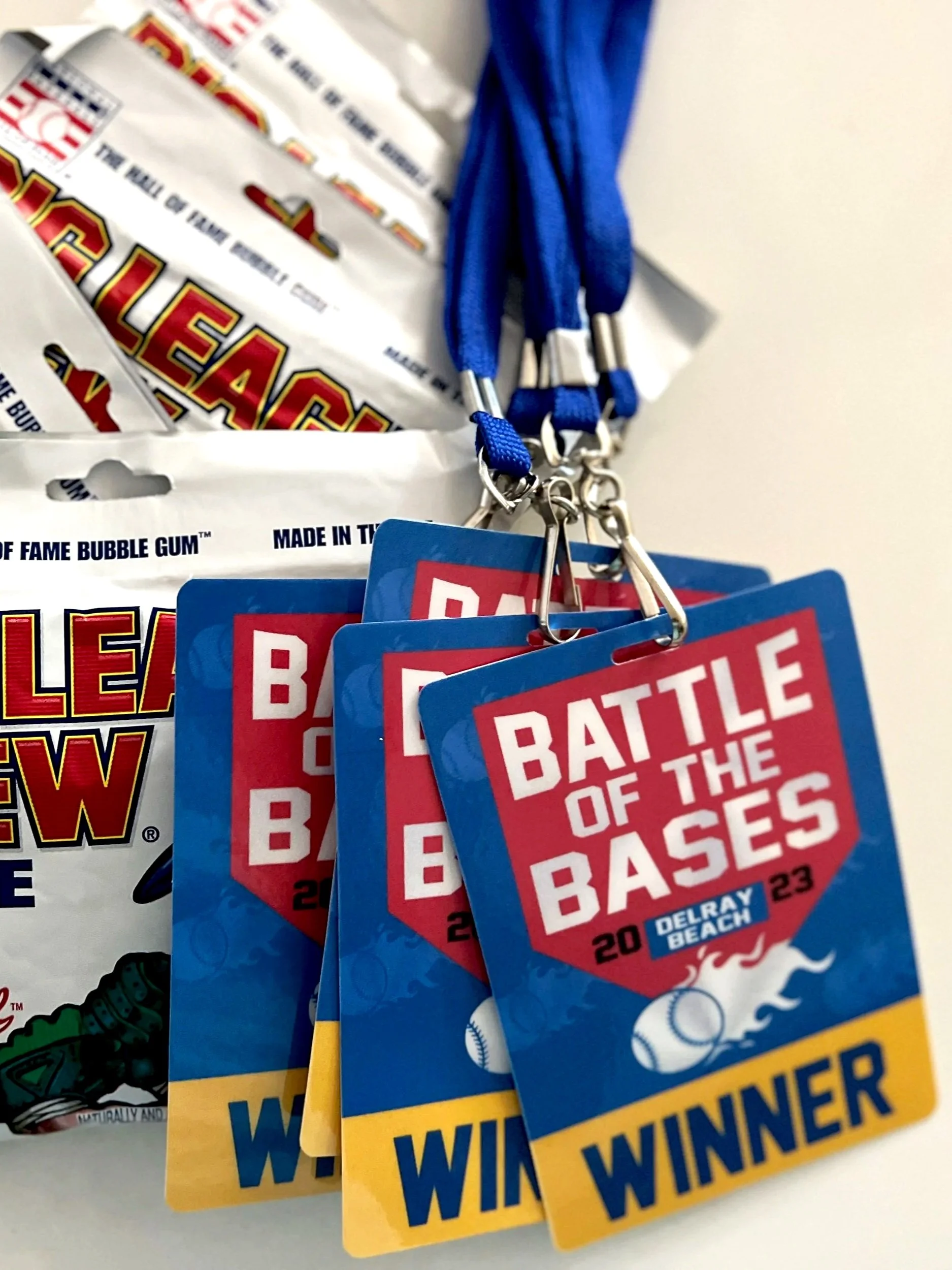 Group of blue rectangular cards with red and white text reading 'Battle of the Bases,' featuring baseballs, with blue lanyards and metal hooks attached, and some brochures in the background.