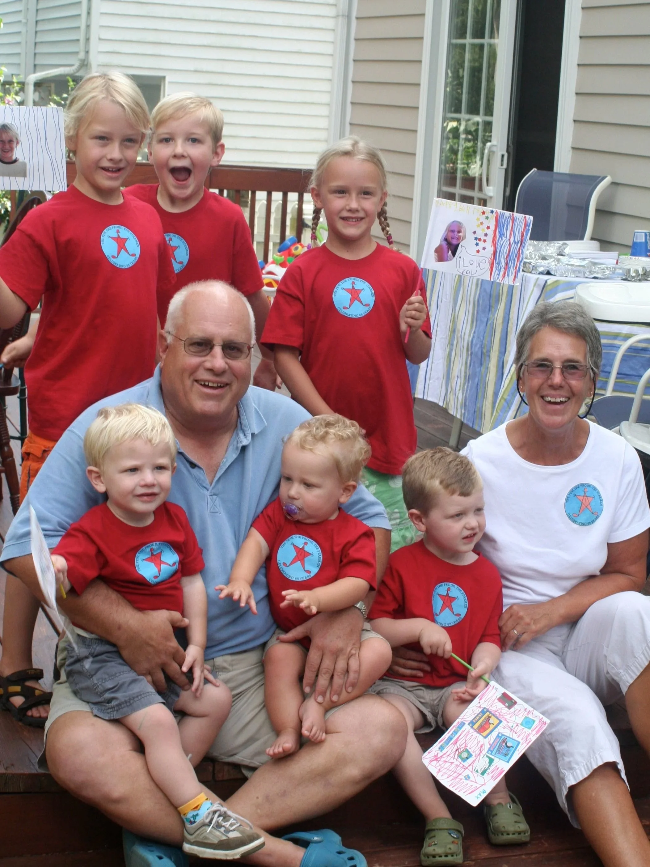 A group of children and adults at a birthday party, with children wearing red shirts with a blue and red star emblem, sitting and standing together outdoors near a table with food and decorations. Some children are holding flags and drawings.