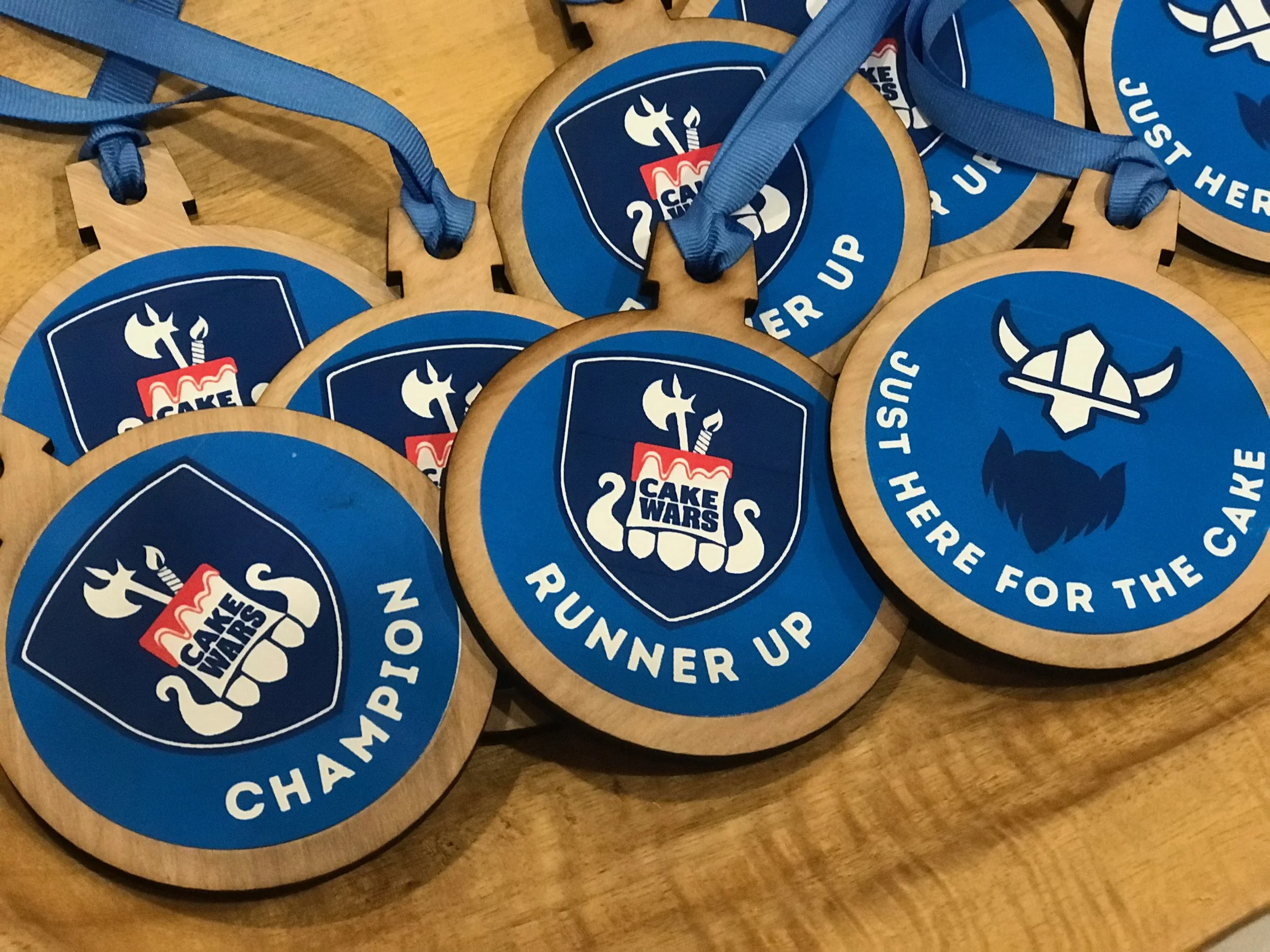 Round wooden medals with blue backgrounds and logos for a cake baking competition titled "Cake Wars" and a Viking helmet with crossed axes, attached to blue ribbons.