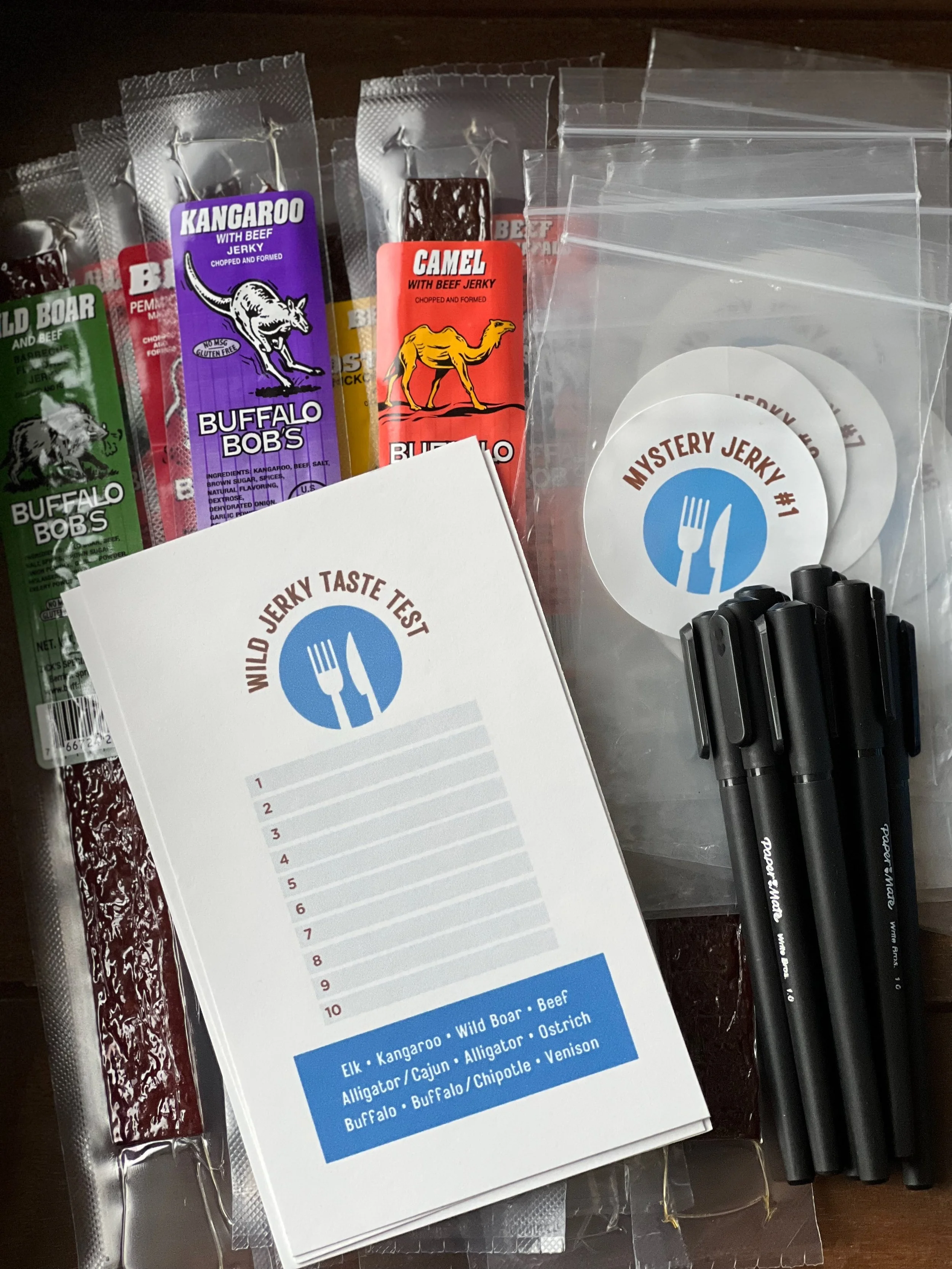 Various packages of beef jerky from Buffalo Bob's, including flavors such as Kangaroo with beef, Camel with beef, Wild Boar with beef, and others, along with a tasting sheet and black pens.