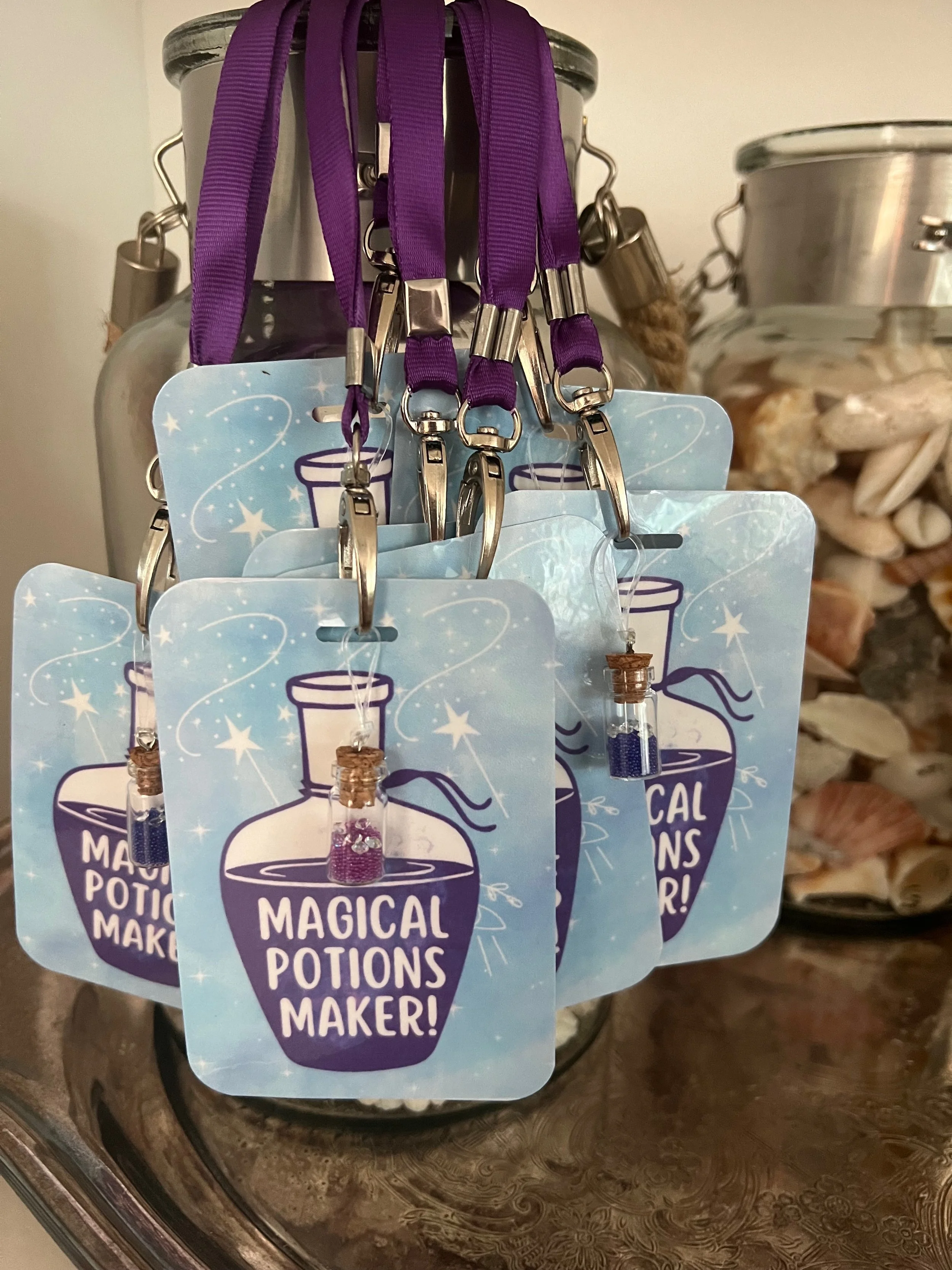 A display of purple lanyards with metal clips hanging from a metallic container, and a stack of cards featuring a magic potion bottle and the words "Magical Potions Maker!".