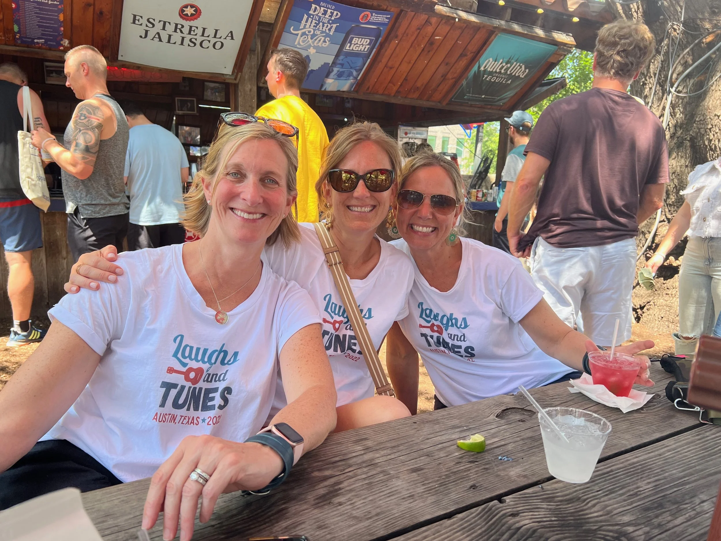 Three women smiling at the camera at an outdoor event, wearing white T-shirts that read 'Laughs and Tunes' with a guitar graphic, seated at a wooden table with drinks, in Austin, Texas.