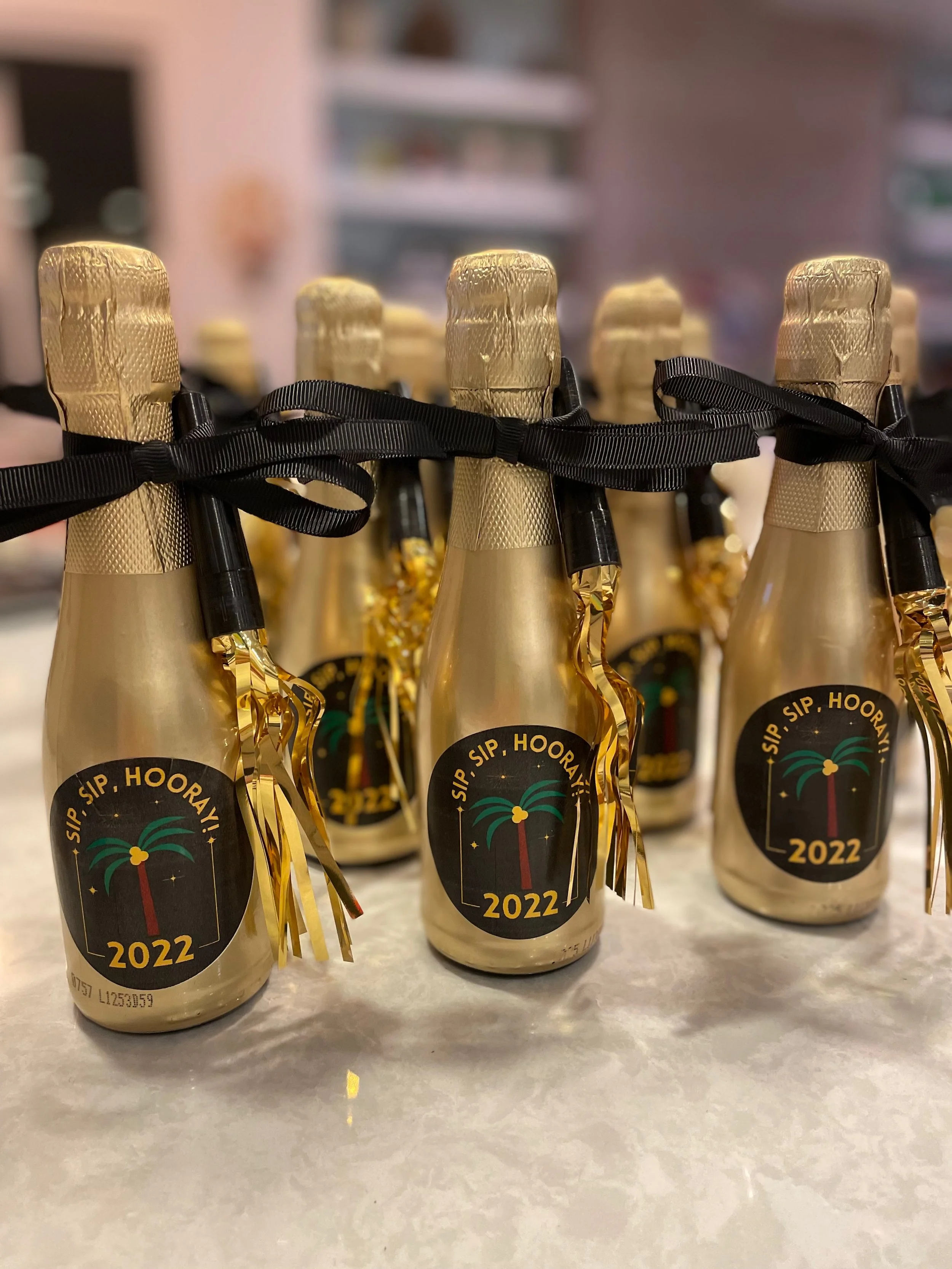 Mini champagne bottles wrapped in gold foil with black ribbons, featuring a holiday-themed label that says 'Sip, Sip, Hooray! 2022'.