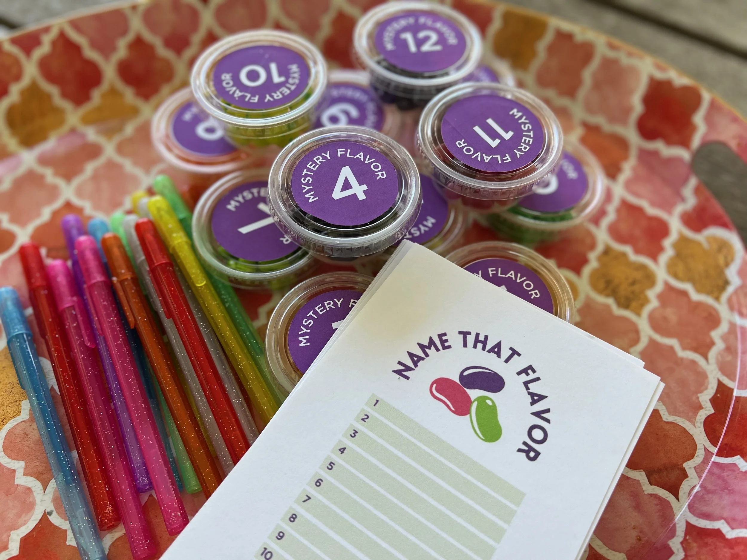 Multiple small containers labeled with purple stickers displaying numbers and mysterious flavors, alongside colorful markers and a partially visible sheet of paper with a flavor testing chart, all placed on a patterned surface.