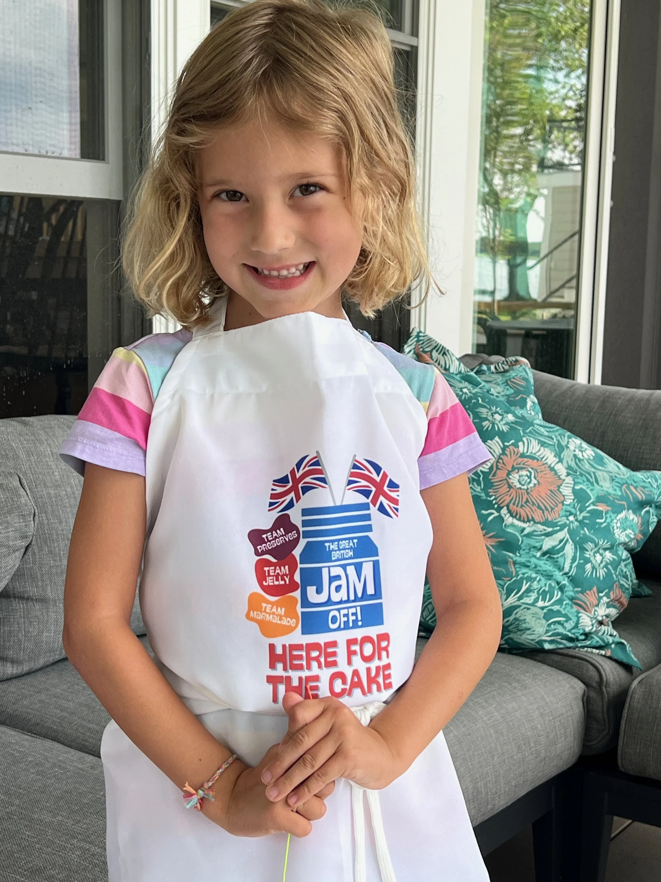 A young girl wearing a white apron with text about jelly, jam, marmalade, and cake, standing indoors near a gray couch and a patterned pillow, with a room featuring large windows and greenery outside in the background.