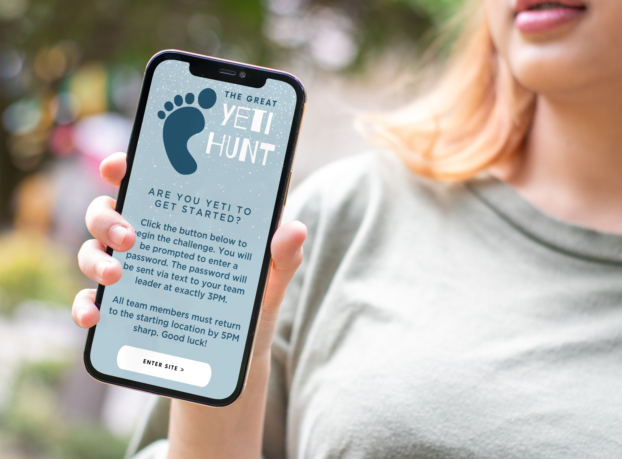 A person holding a smartphone displaying a website titled 'The Great Yeti Hunt' with instructions to click a button to begin a challenge, with a subtitle about entering a site by 3PM and returning to the starting location by 5PM.