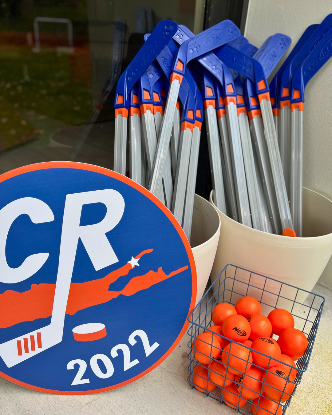 A pile of blue and silver telescoping q-tips leaning against a beige wall, a blue and orange campaign sign with the text 'CR 2022' and an image of Texas with a star, and a metal basket filled with orange ping pong balls.