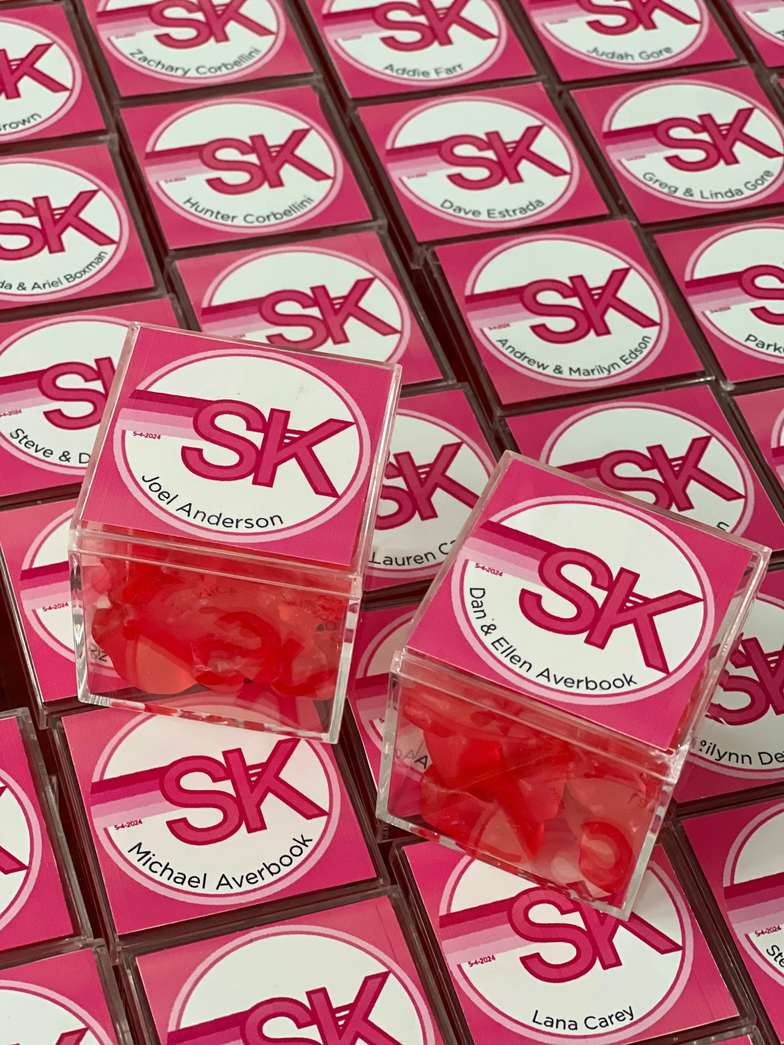 Multiple pink stickers with white circles and red letters spelling 'SK', each with a different name underneath, displayed on a wall. Some of the names include Joel Anderson, Mike & Ellen, Lauren Carey, Dan & Ellen, Greg & Linda, and others.