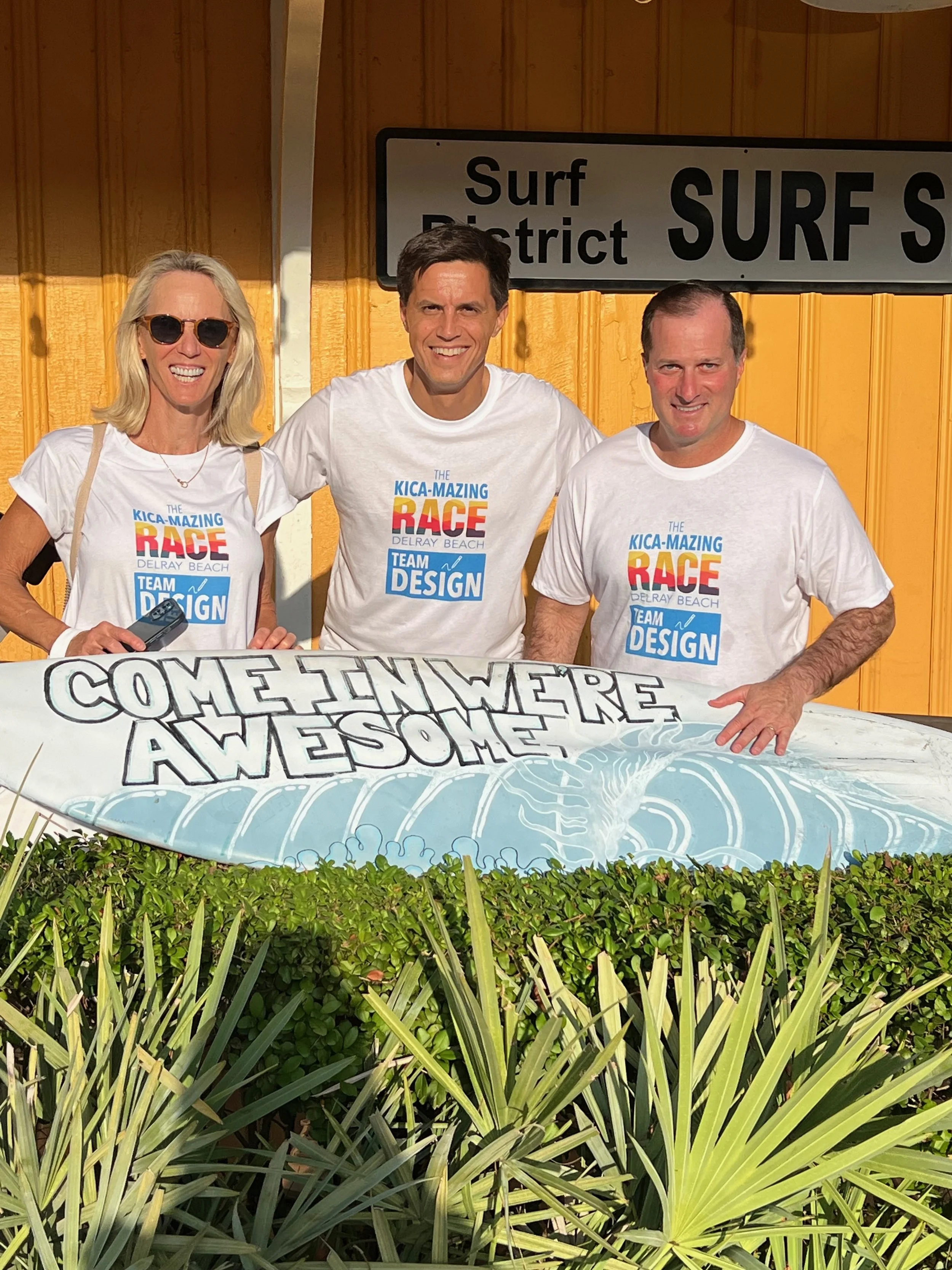 Three people standing behind a sign that says "Come in we're awesome" with a background of yellow wooden wall and green plants in front.