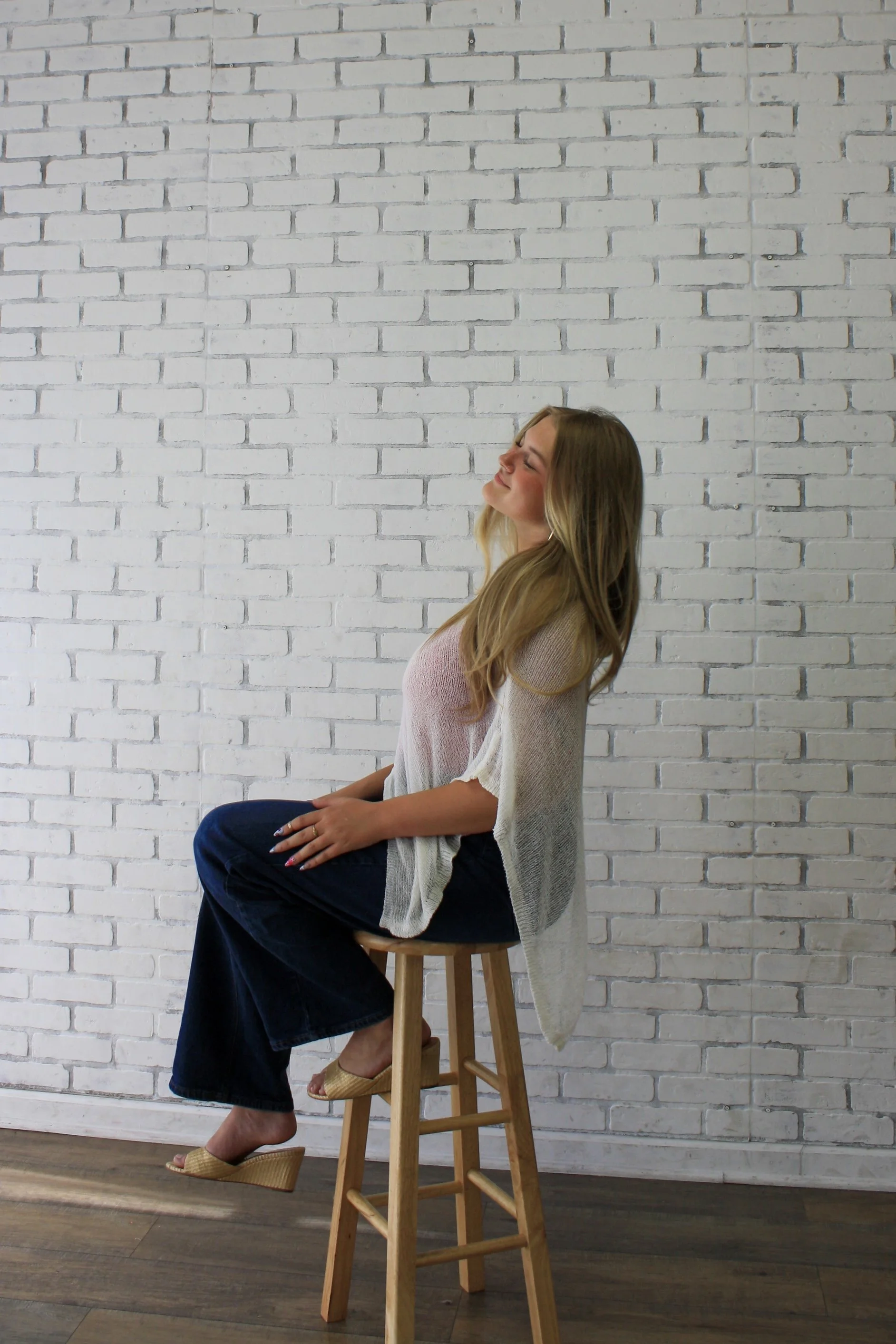 A woman with long blonde hair sitting on a stool, smiling with eyes closed, against a white brick wall, wearing a beige top, dark pants, and beige wedge sandals, with a beige cardigan draped over her shoulders.