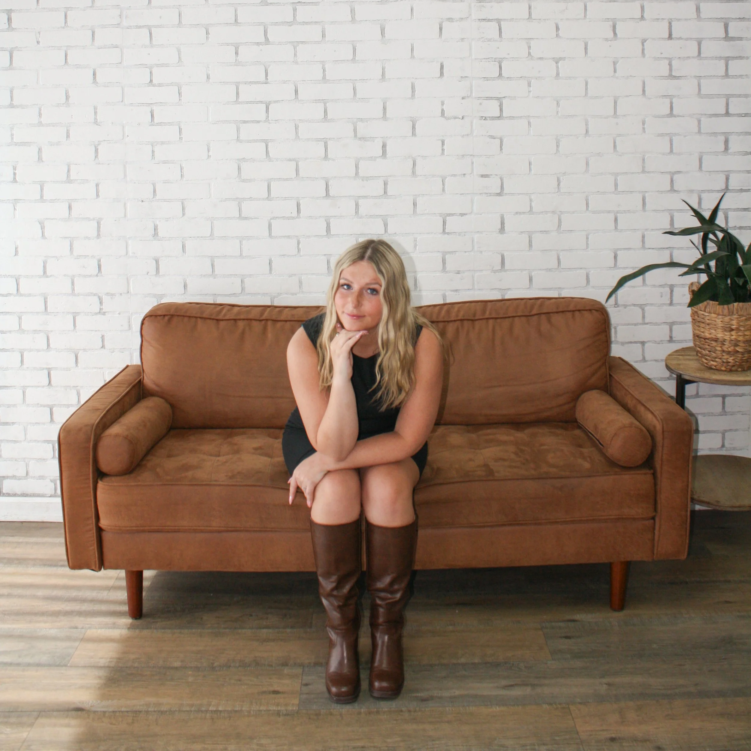 A woman sitting on a brown sofa with a white brick wall in the background, wearing a black top and brown knee-high boots, with a potted plant nearby.