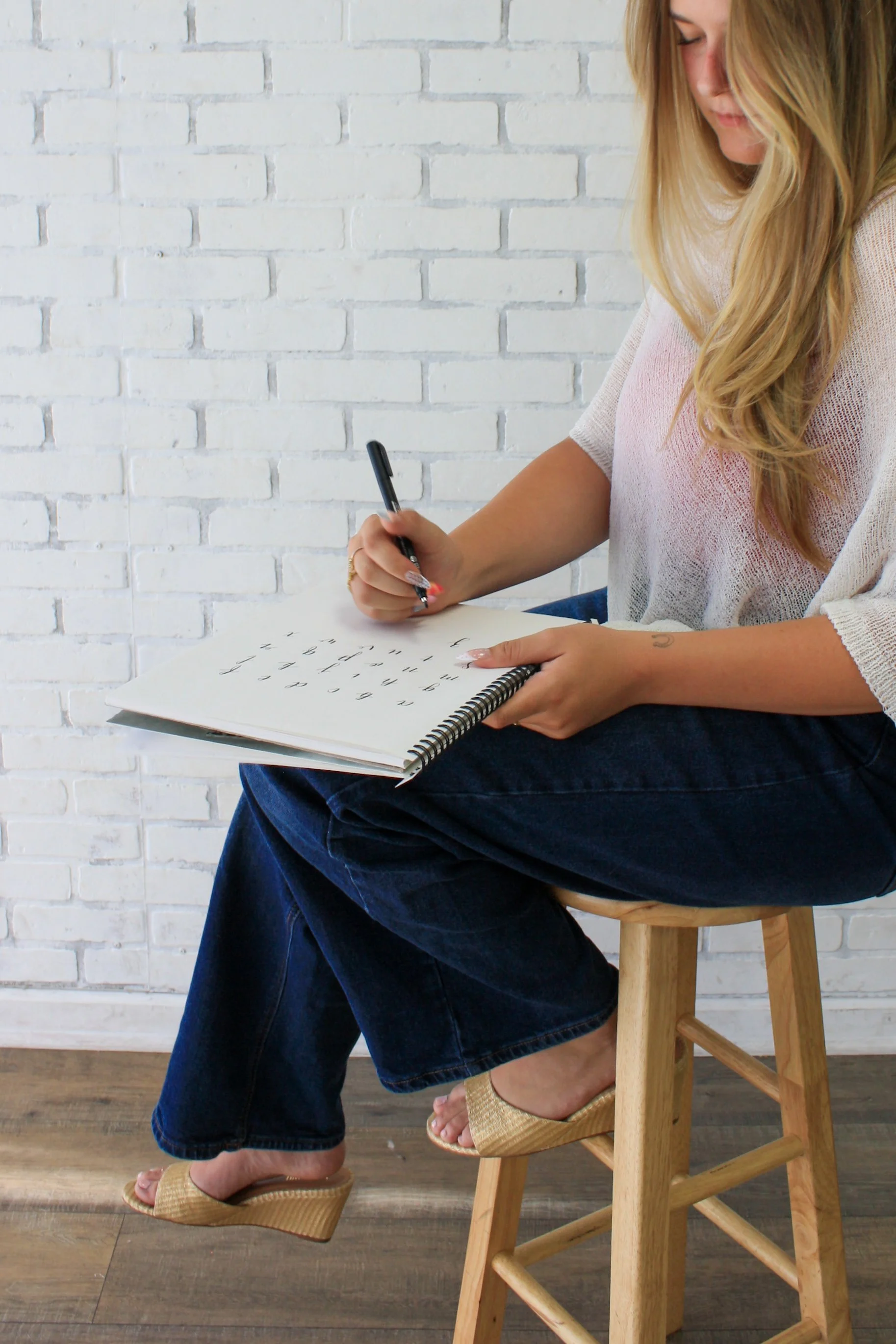 A woman sitting on a wooden stool, writing in a spiral notebook with a black pen, wearing a light pink top, dark blue jeans, and tan wedge sandals, against a white brick wall background.
