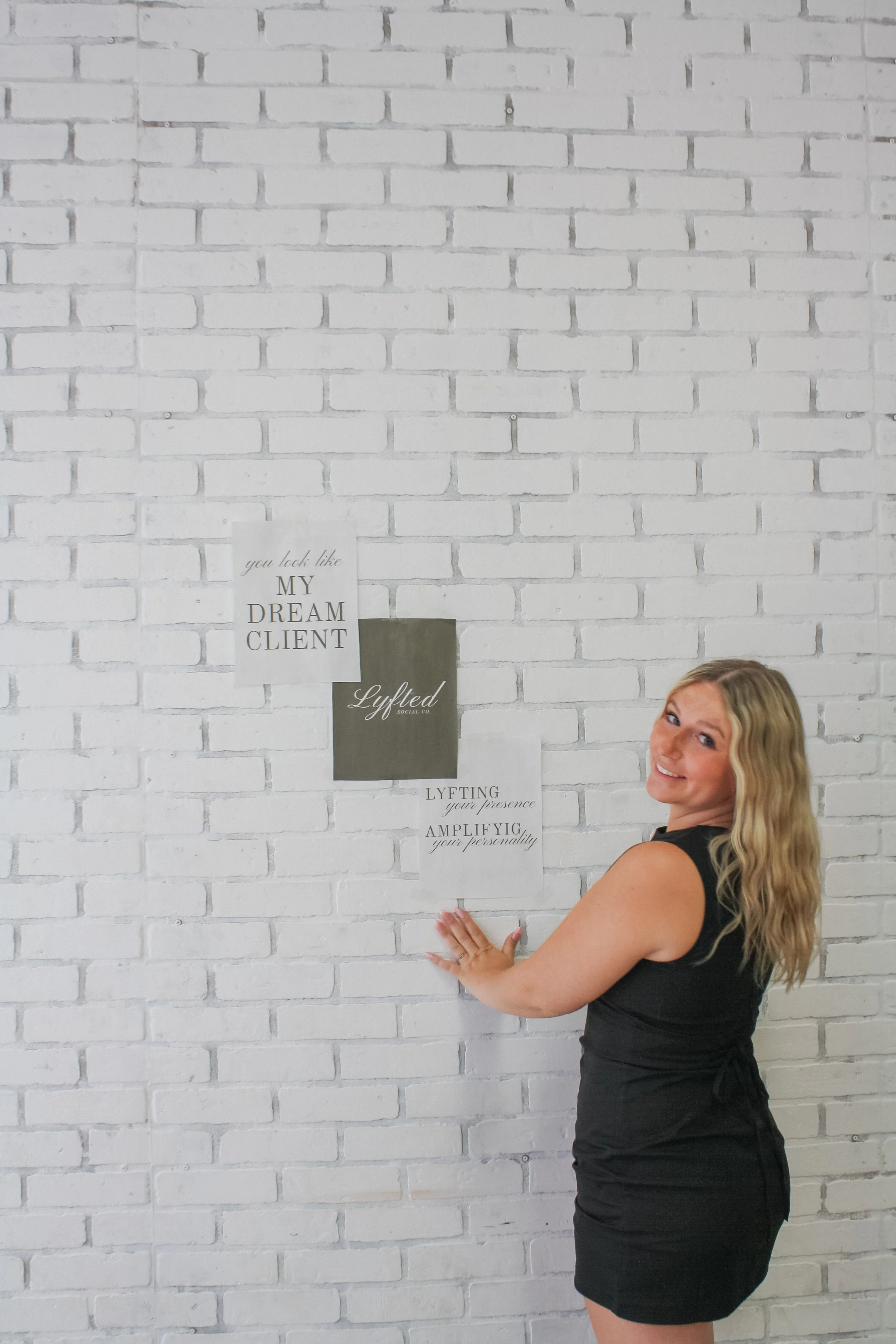 A woman with long blonde hair wearing a black sleeveless dress is standing in front of a white brick wall with motivational posters, smiling at the camera.