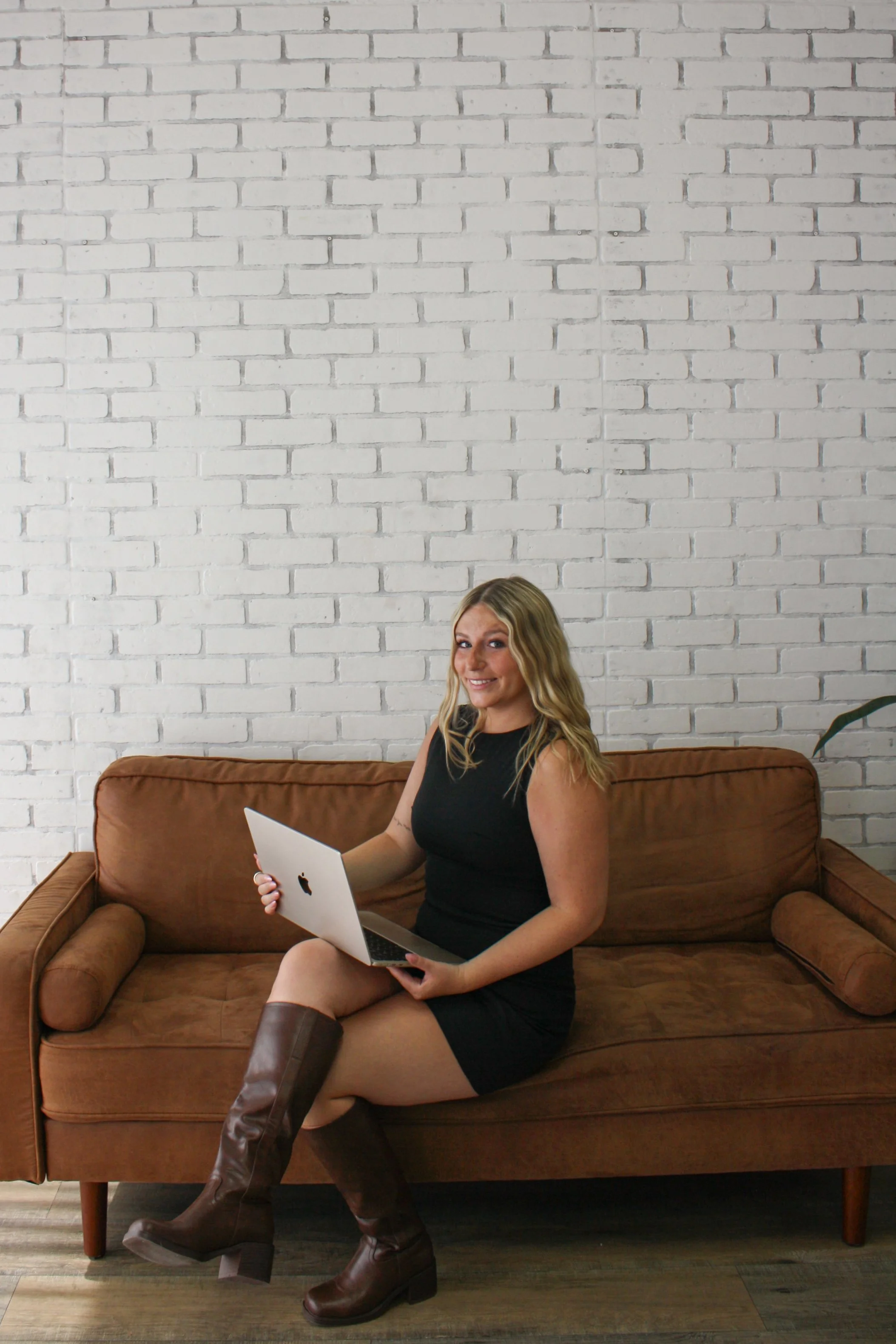 A woman sitting on a brown sofa, holding a silver laptop, wearing a black sleeveless dress and tall brown boots, with long blonde hair, smiling, in front of a white brick wall.