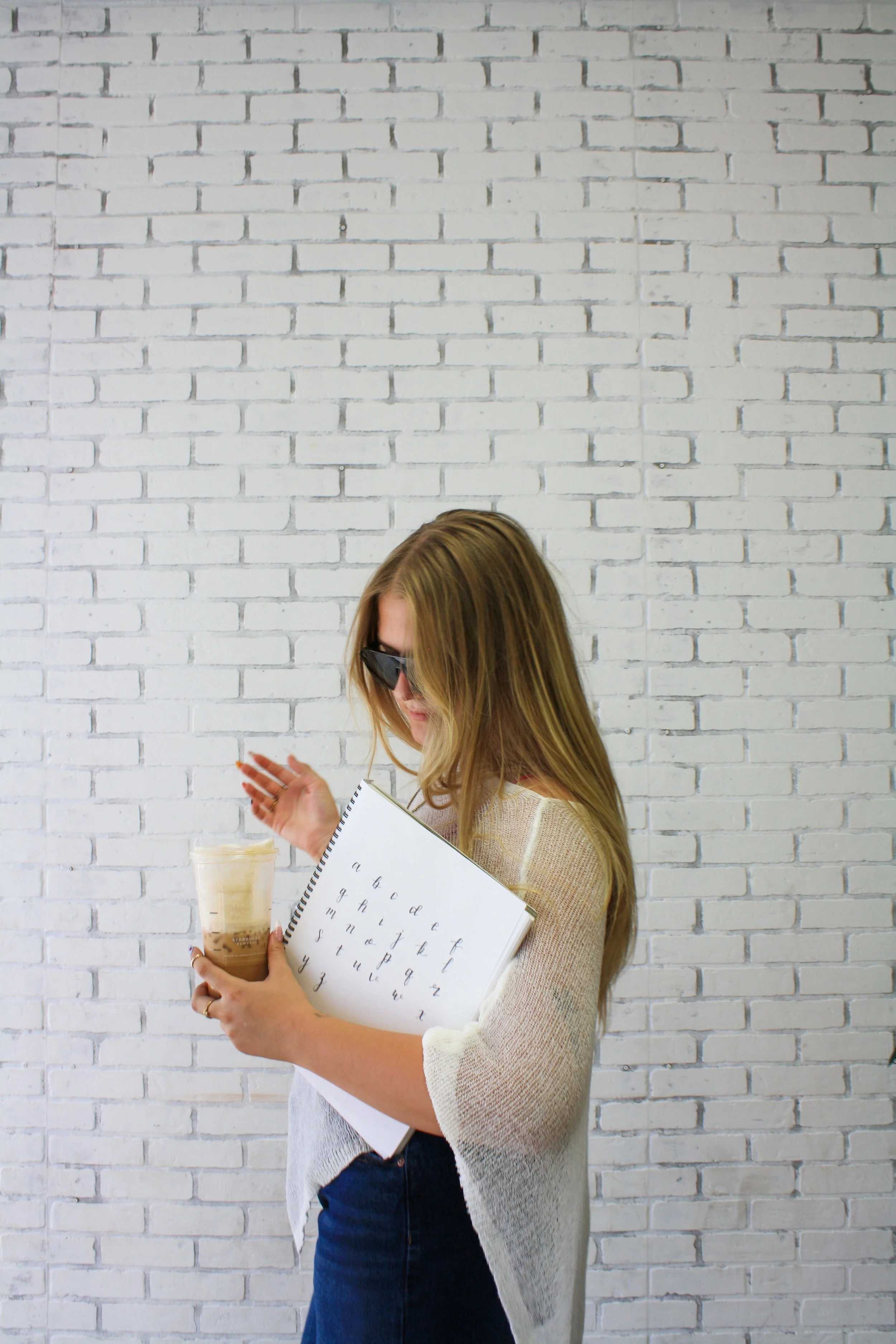 Woman with long blonde hair and glasses, wearing a beige cardigan over a white shirt, holding a Starbucks iced coffee in one hand and a notebook with alphabet practice in the other, standing against a white brick wall.