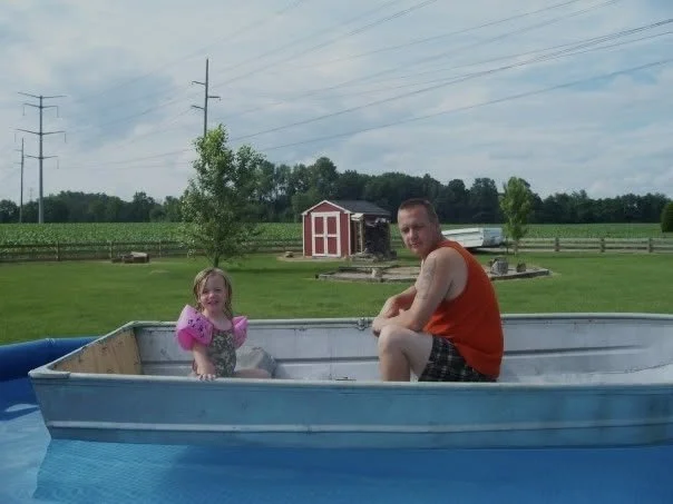A man and a young girl in a small boat on a pond or lake, with a farm landscape in the background including barns, trees, and power lines.