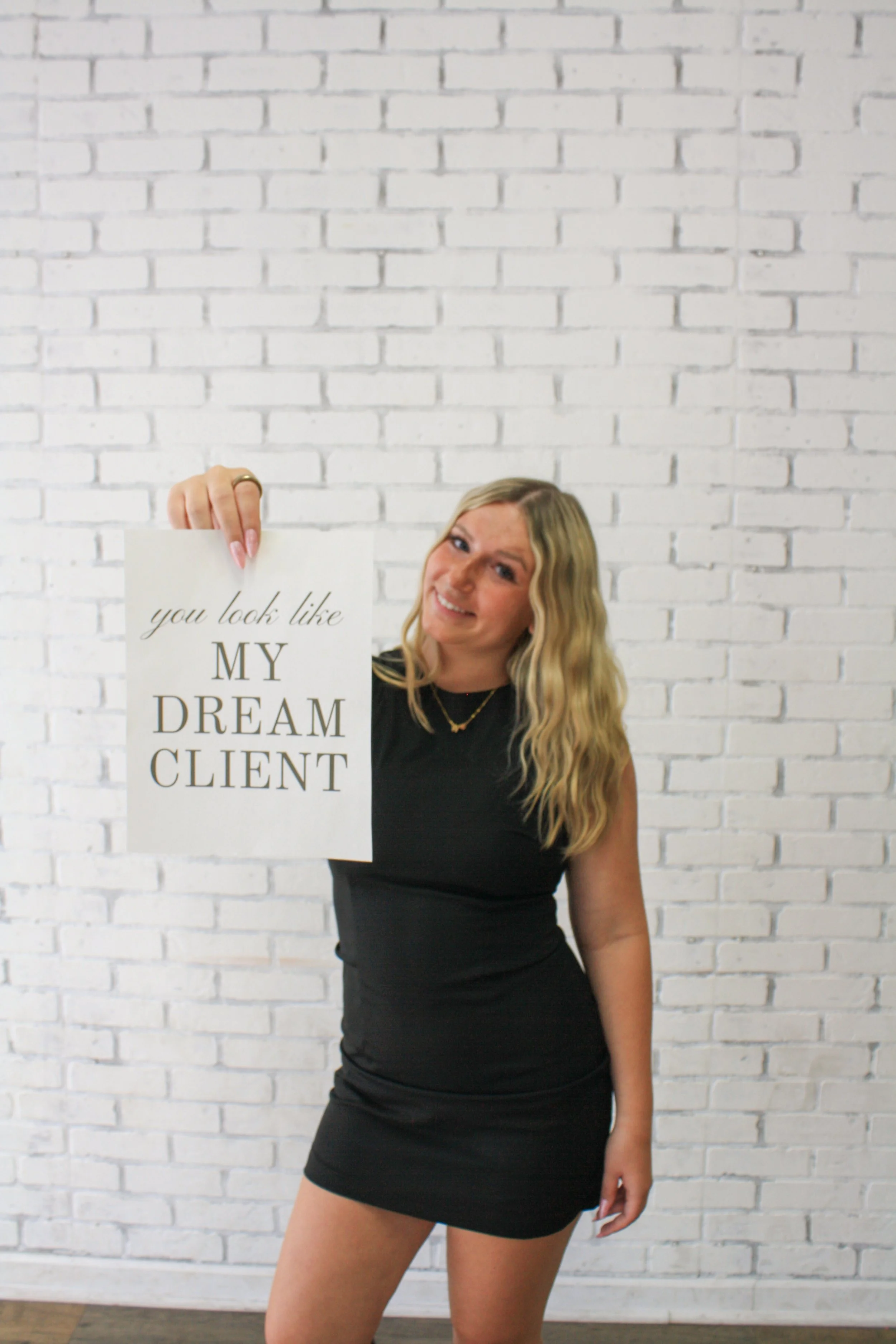Woman with long blonde hair wearing a black dress, smiling, holding a sign that says "you look like MY DREAM CLIENT" against a white brick wall background.