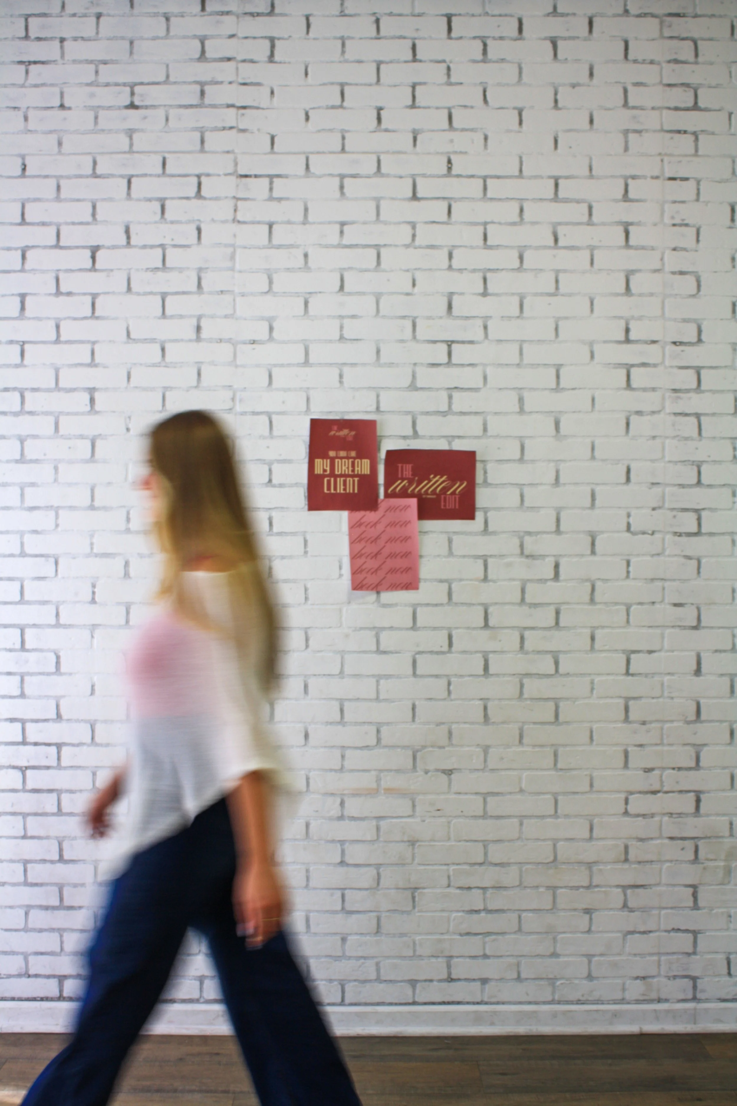 A person walking past a white brick wall with red posters attached. The posters have text, but it is difficult to read due to motion blur.