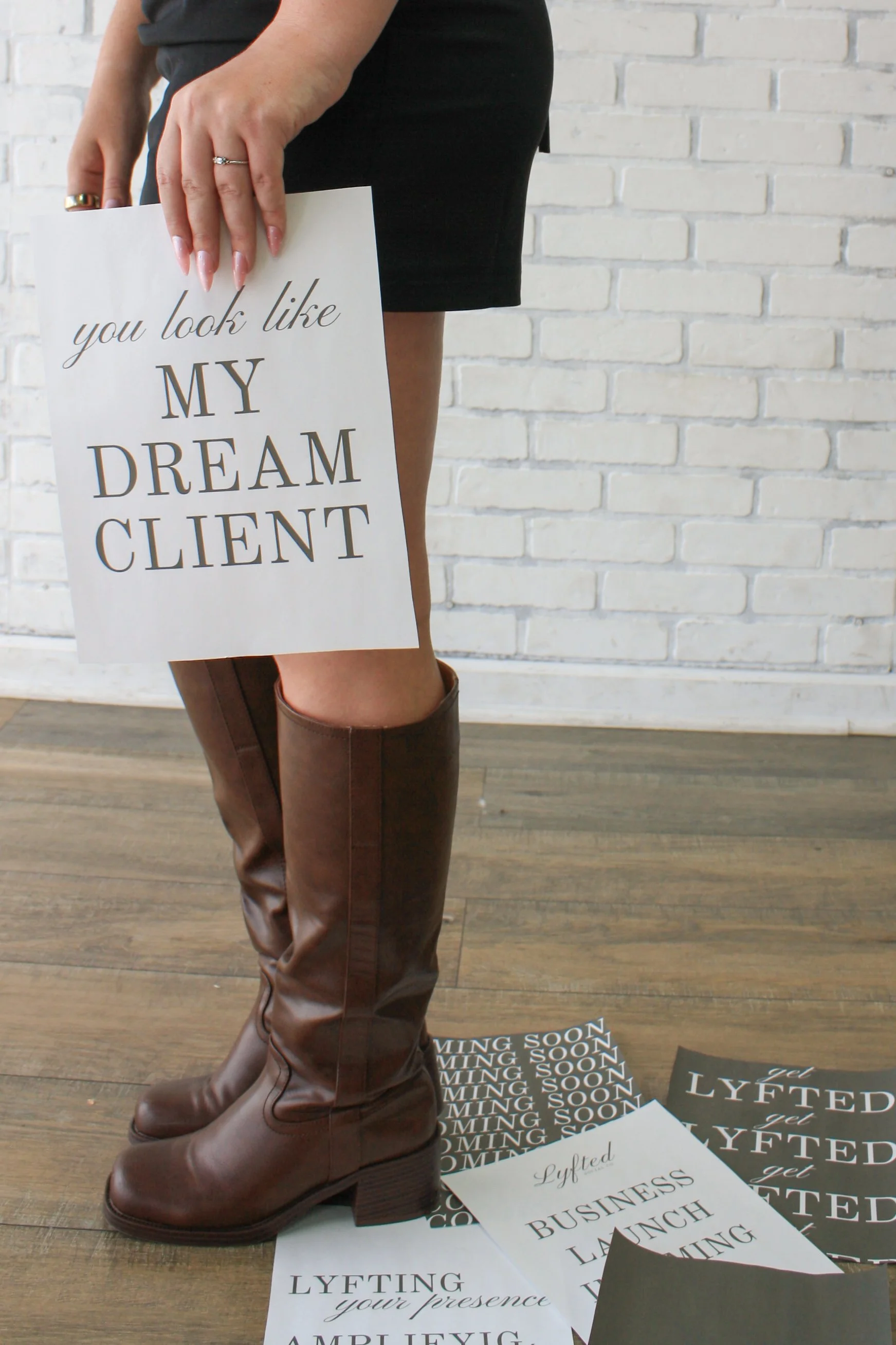 A person holding a sign that says 'you look like MY DREAM CLIENT'. The person is wearing brown boots and a black dress, standing on a wooden floor with printed signs spread around.