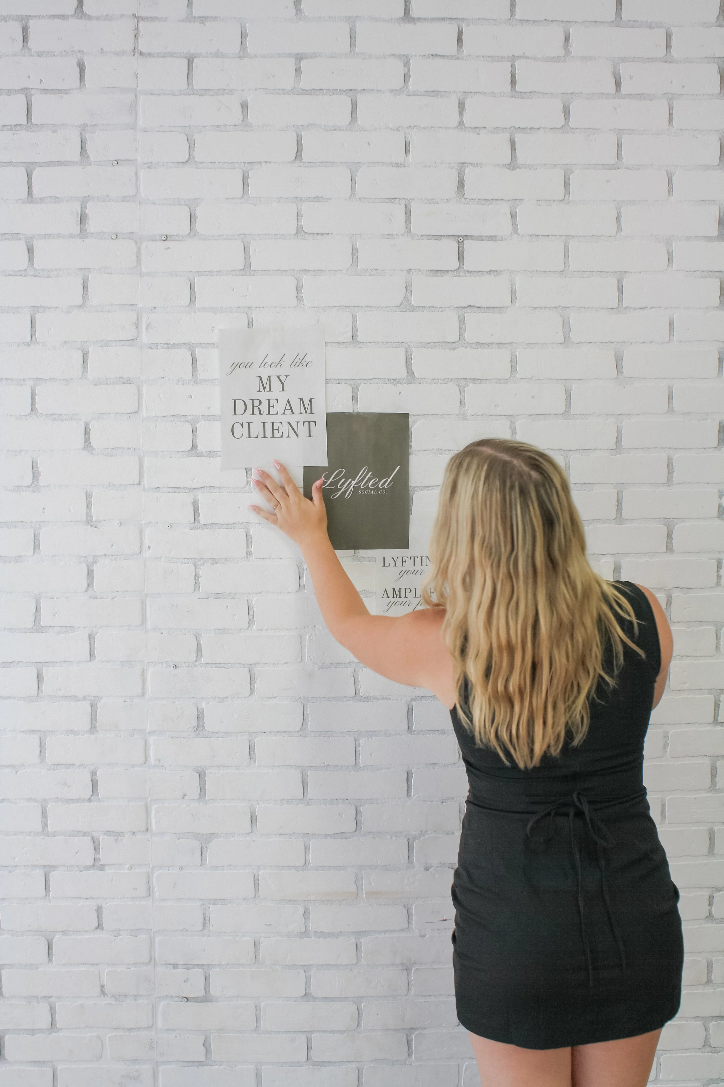 A woman with long blonde hair wearing a black dress, standing against a white brick wall, hanging a sign that says 'you look like MY DREAM CLIENT'.