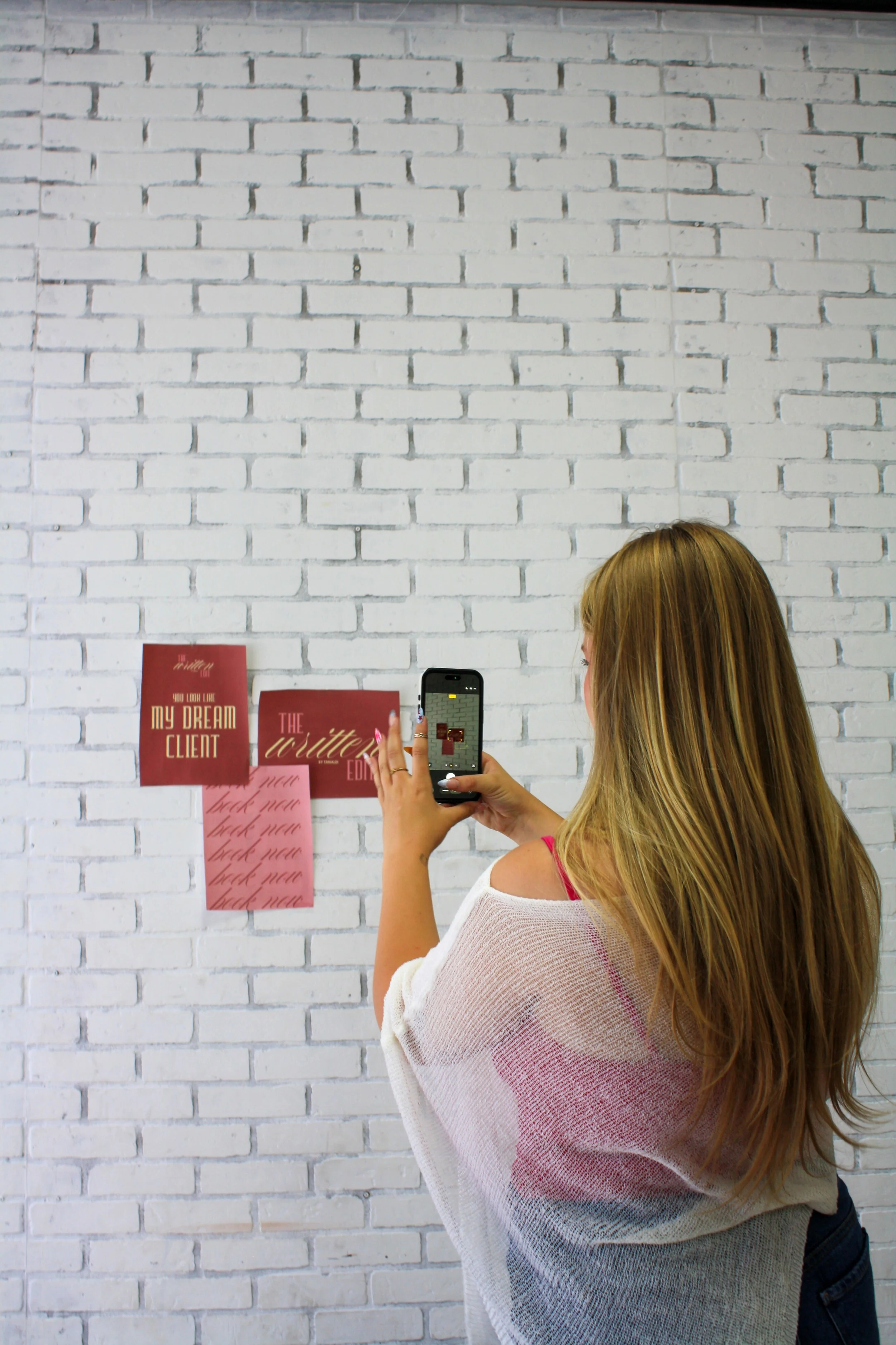 A woman with long, blonde hair taking a photo with her phone in front of a white brick wall. The wall has red and pink paper notes taped to it, with some of the notes reading "My Dream Client" and "The Writer."