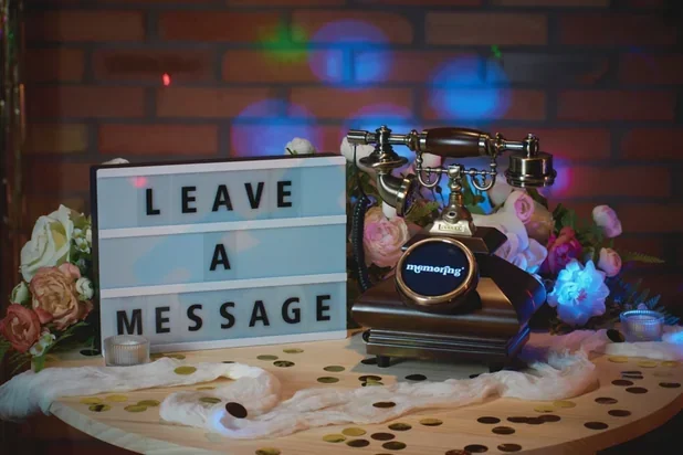 Decorative table with a vintage rotary phone, flowers, a lightbox with 'Leave a Message,' and confetti on a table with a brick wall background.