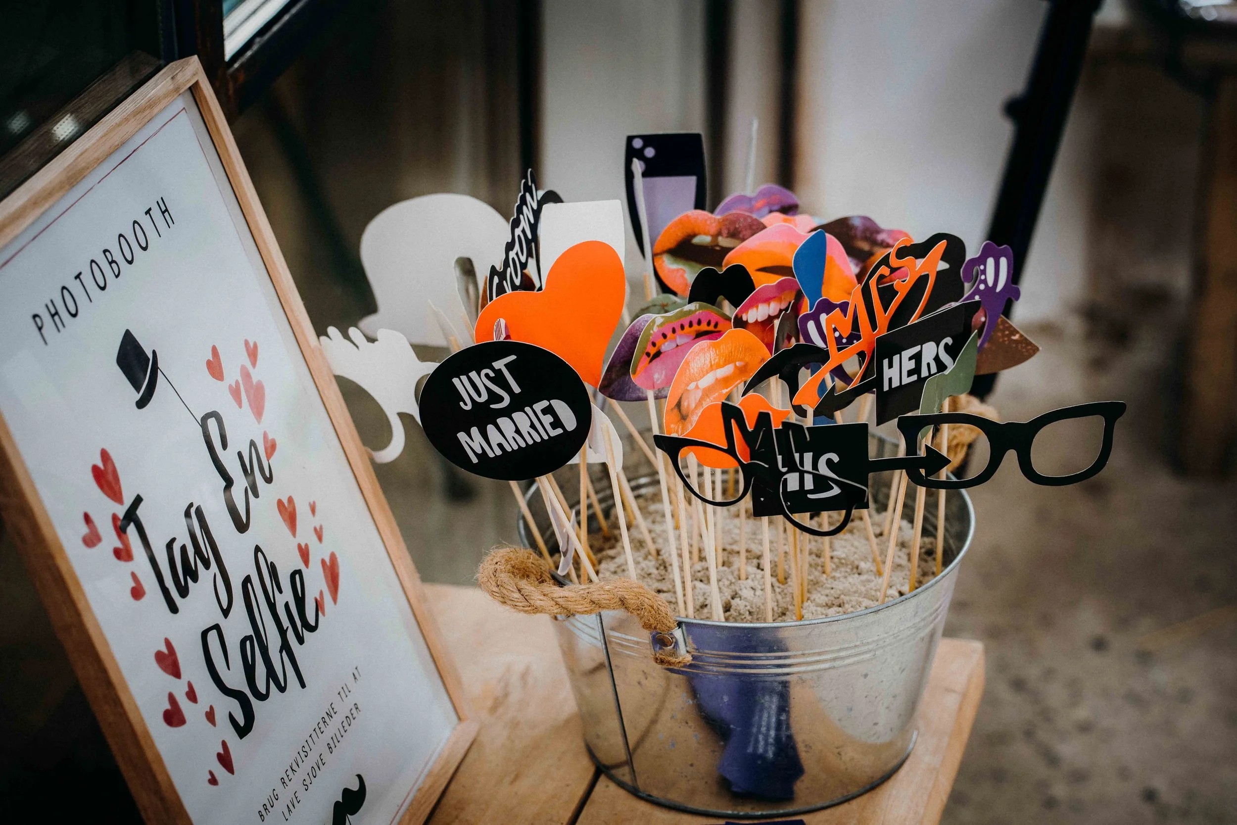 Decorative photo booth props with lips, hearts, and signs, arranged in a metal bucket beside a sign with red hearts and text, at a wedding or event.