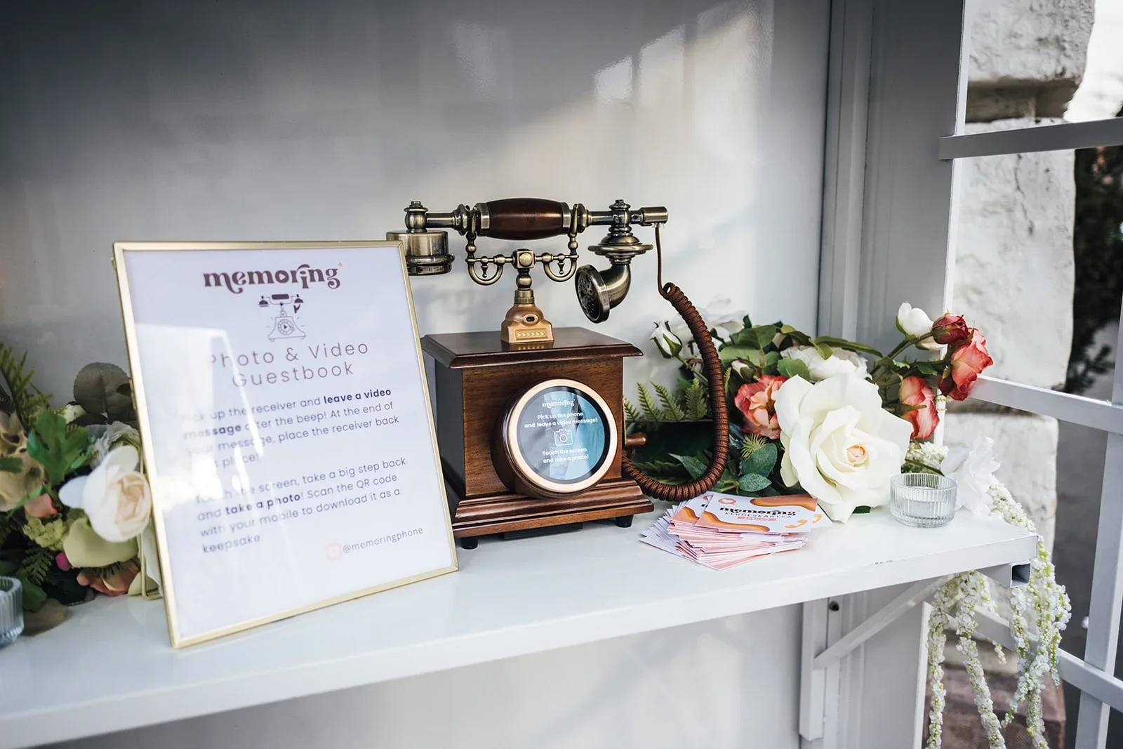 A floral arrangement with white and pink flowers next to a vintage phone on a wooden stand, digital sign with instructions for a photo and video guestbook, and small stacks of paper on a white table.