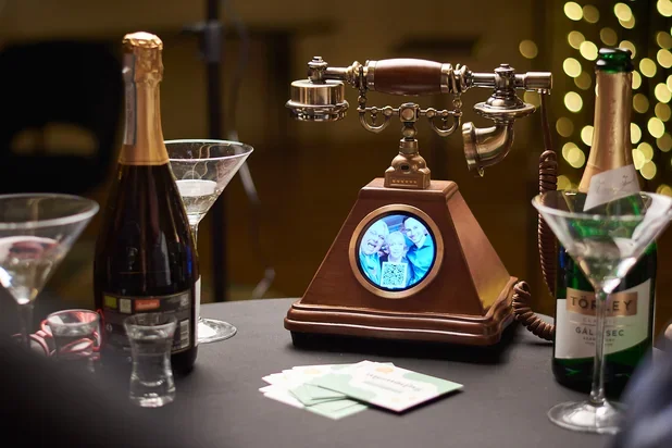Vintage telephone placed on a table surrounded by bottles of wine, cocktail glasses, and money, with blurred festive lights in the background.