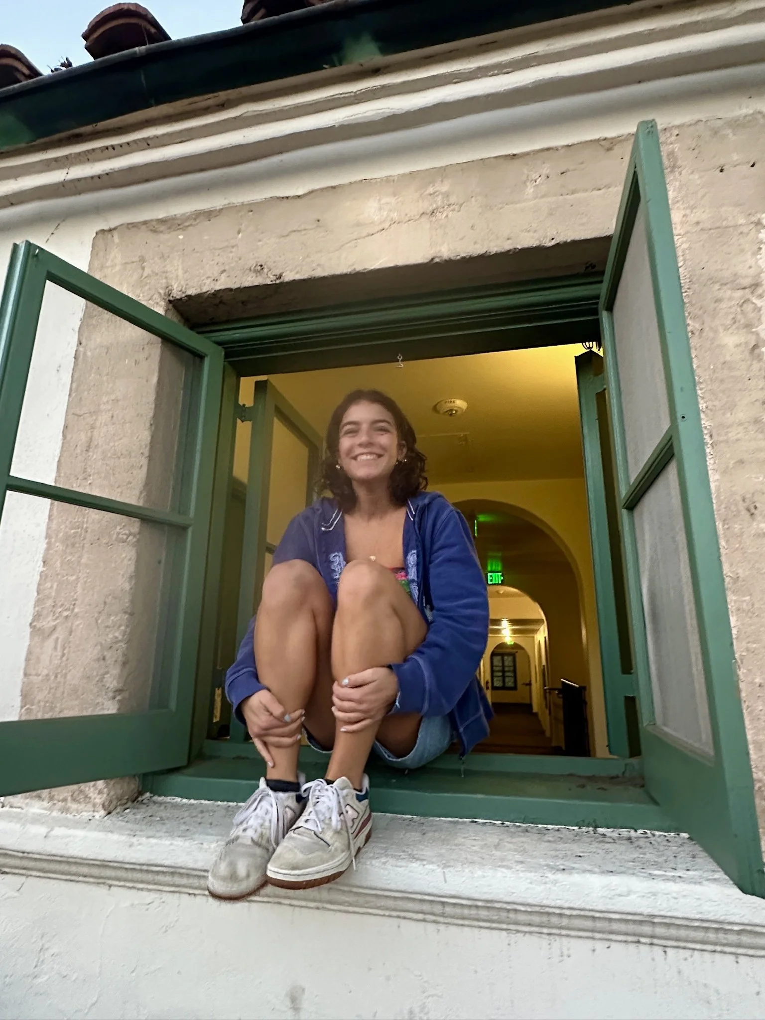 A smiling woman is sitting on a windowsill with her legs tucked in and arms wrapped around her knees. She is wearing a blue jacket, dark shorts, and sneakers. The window has green frames, and the interior of the building is lit with yellow light. The exterior background shows a cream-colored wall and a tiled roof.