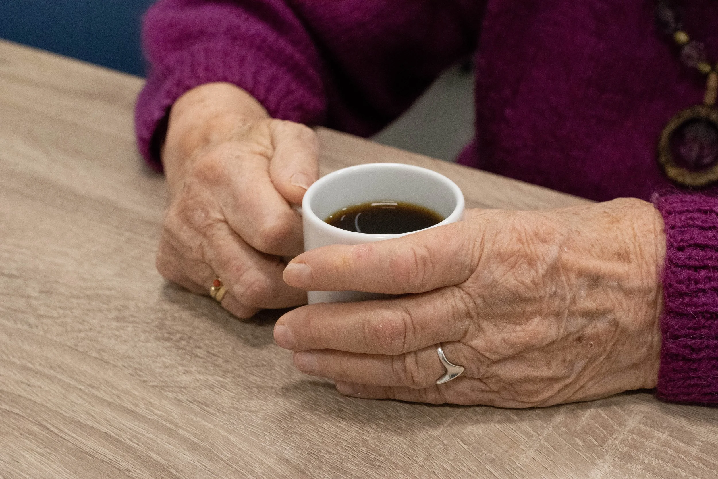 De afbeelding toont de handen van een persoon die een witte kop met koffie vasthoudt, zittend aan een houten tafel.