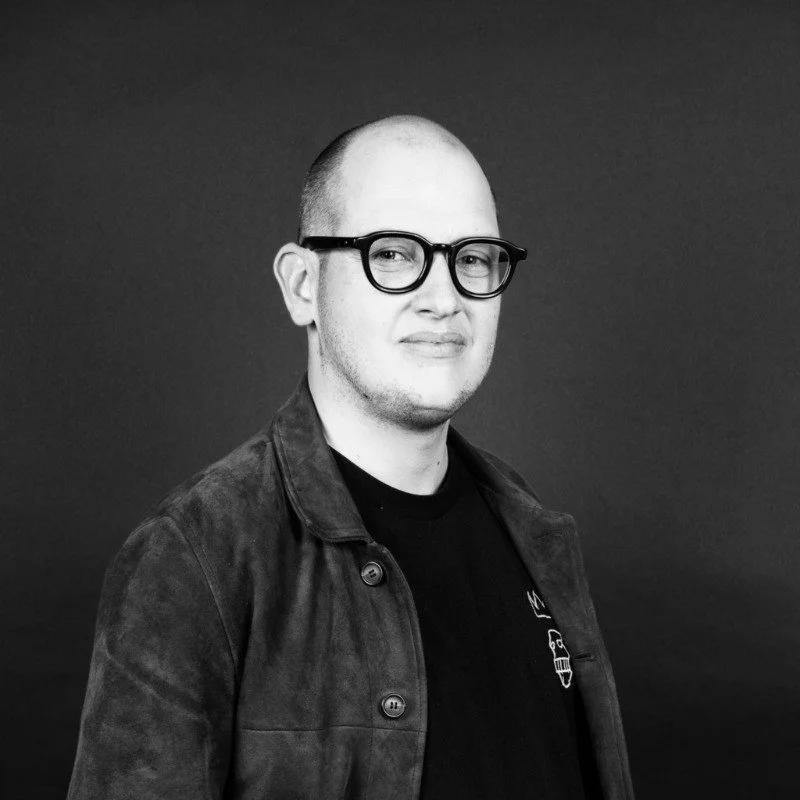 A man with glasses and a shaved head wearing a dark jacket and shirt, standing against a plain dark background.