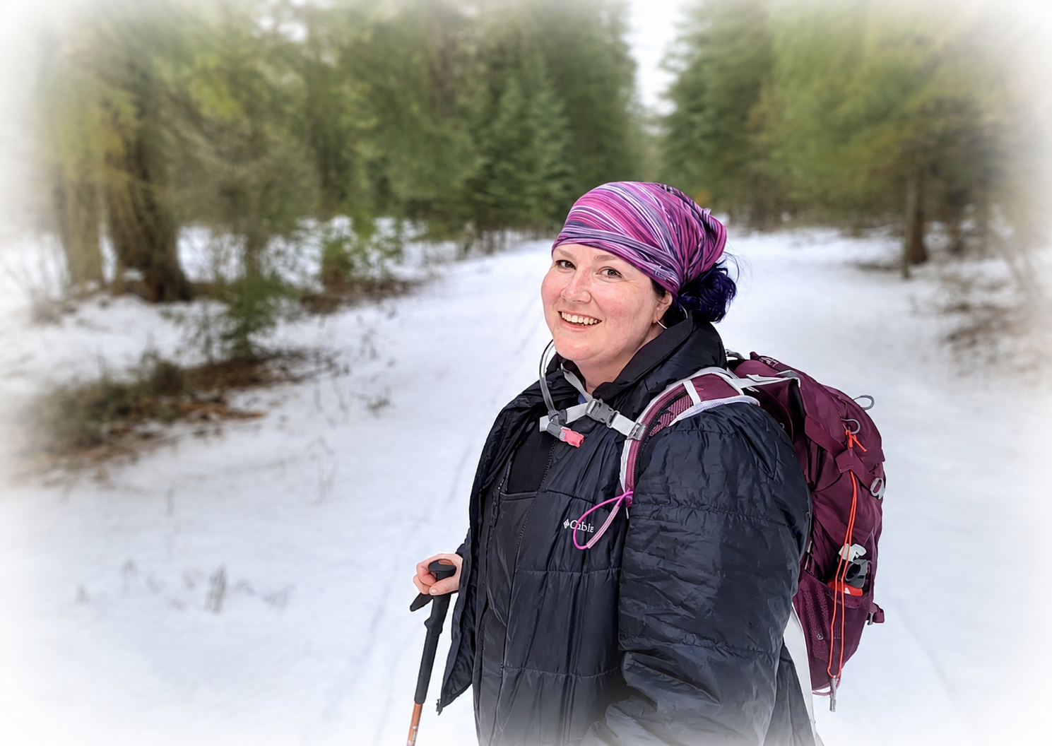 Smiling woman wearing a purple headscarf and black jacket hiking through snow-covered forest with backpack and walking poles.