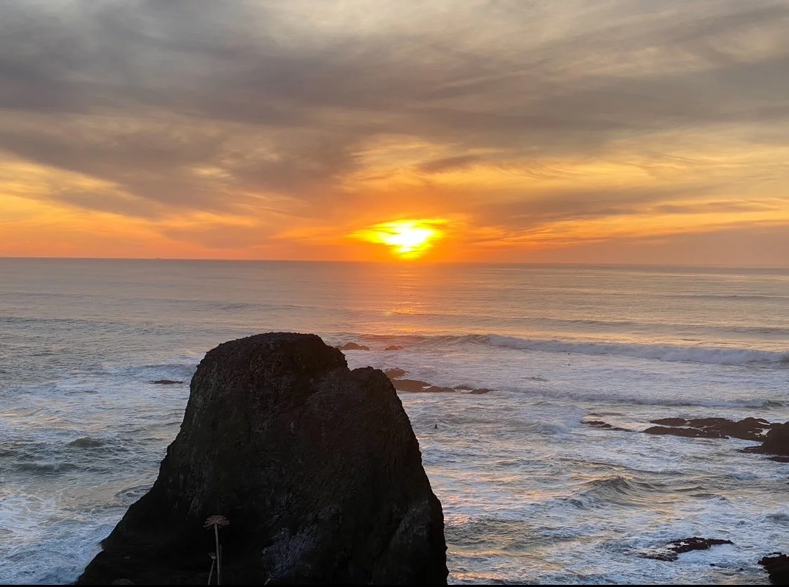Sun setting over the ocean with waves crashing against rocks in the foreground.