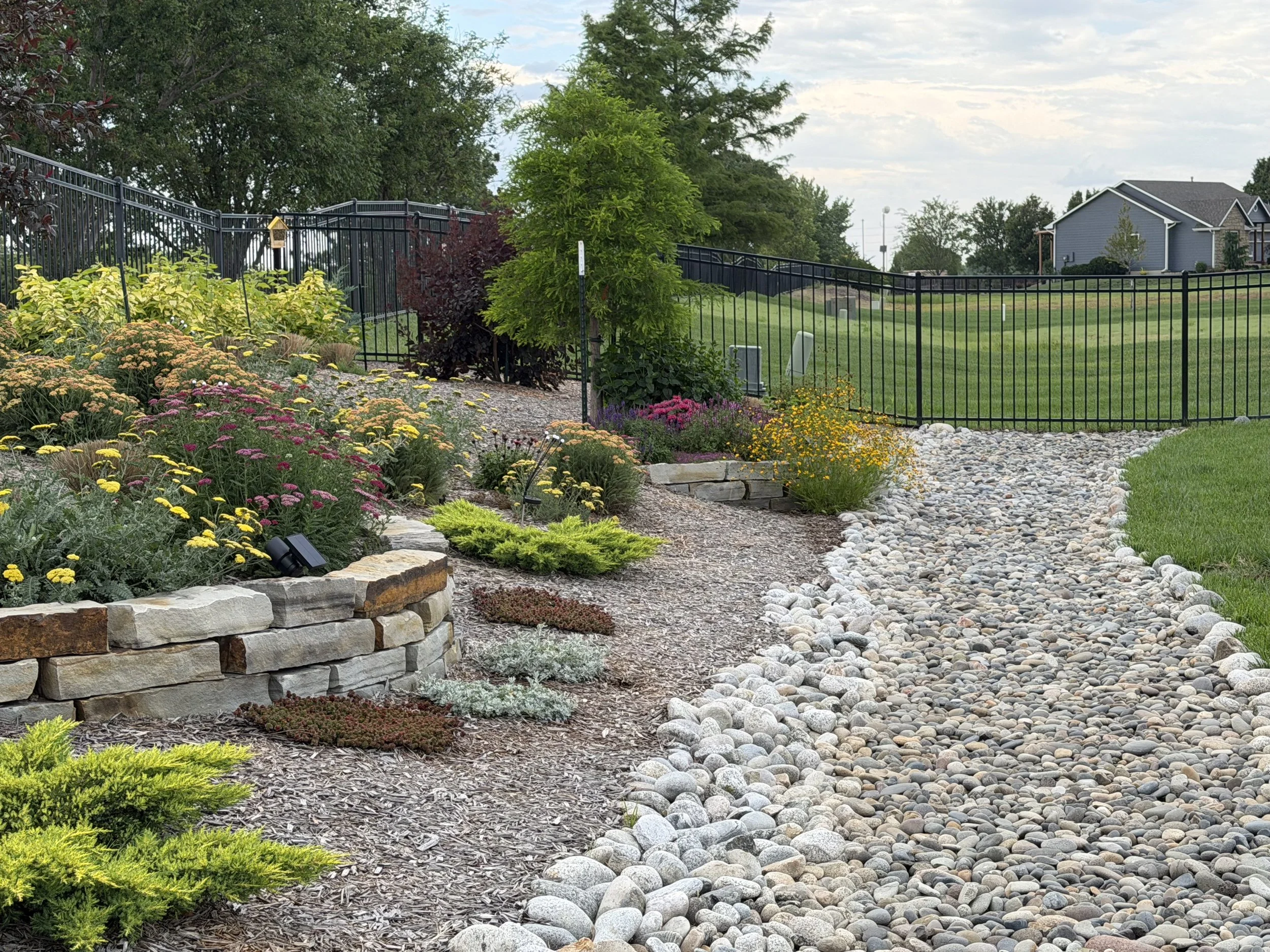 A landscaped yard with flowering plants, a stone retaining wall, a gravel pathway, a black metal fence, and a house with a gray exterior in the background.