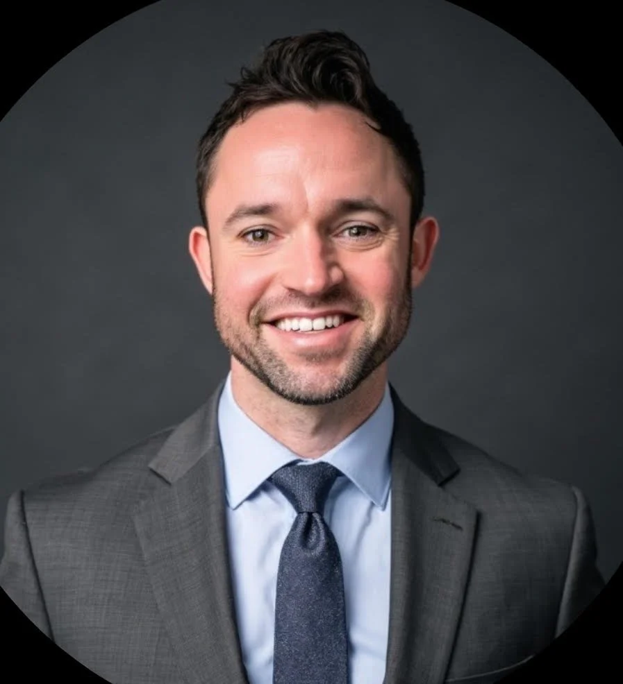Headshot of a smiling man in a gray suit, light blue shirt, and dark tie, with dark hair and a beard, against a dark gray background.