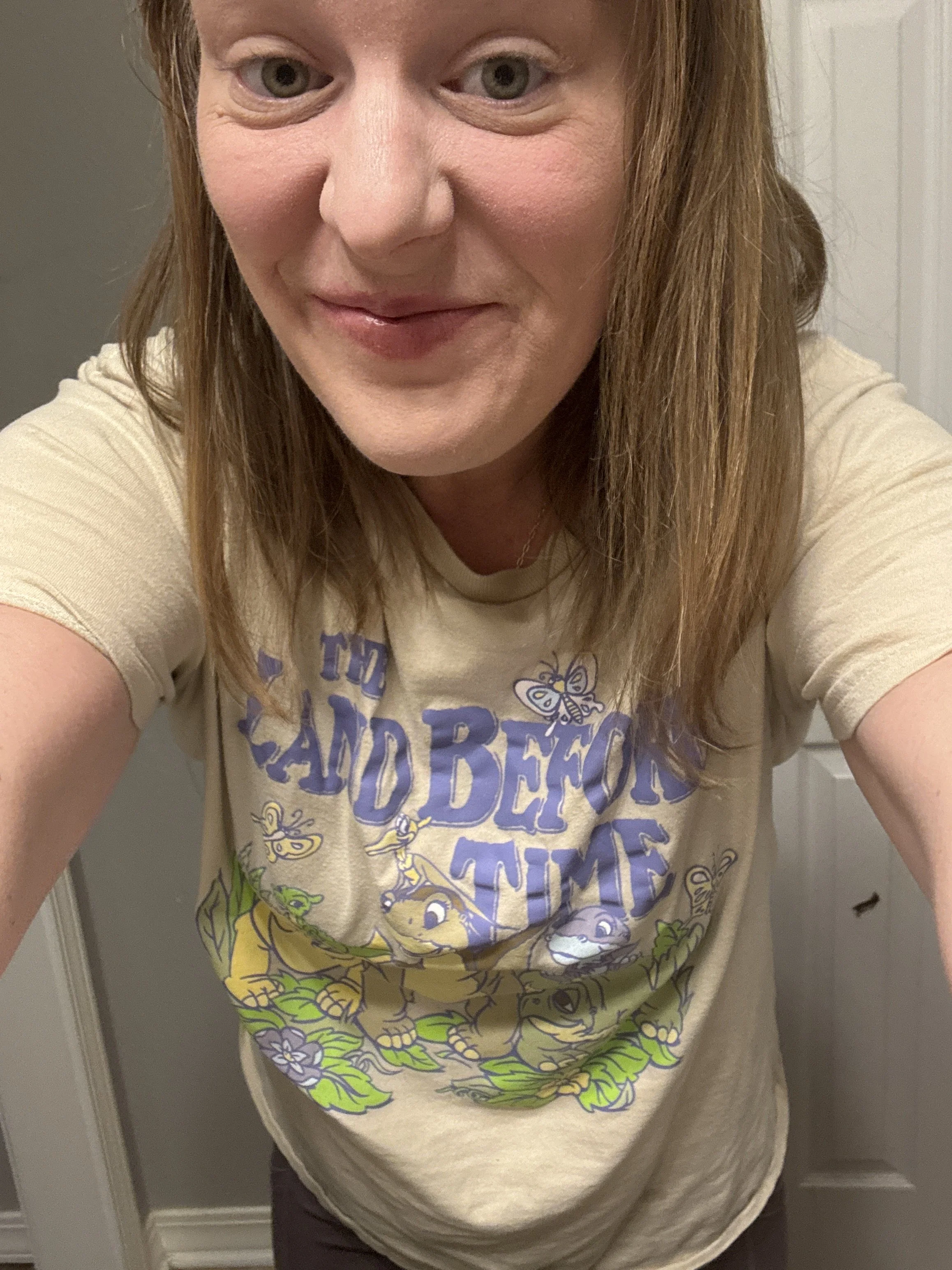 A woman with long reddish hair and greenish-gray eyes taking a selfie in front of a mirror, wearing a beige T-shirt with colorful cartoon characters and text.