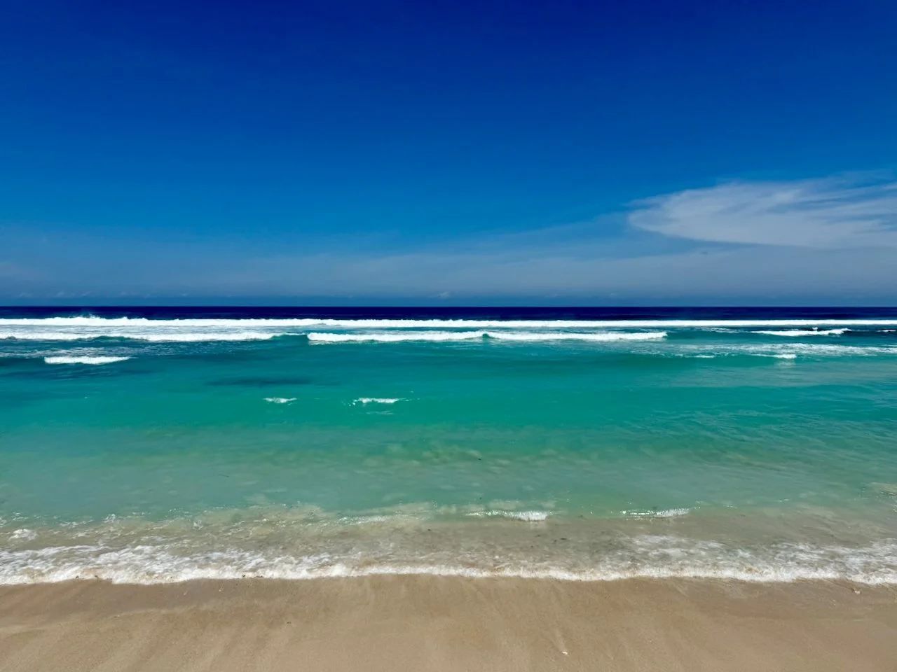 Sunny beach with clear turquoise water, gentle waves, and a blue sky with a few clouds.