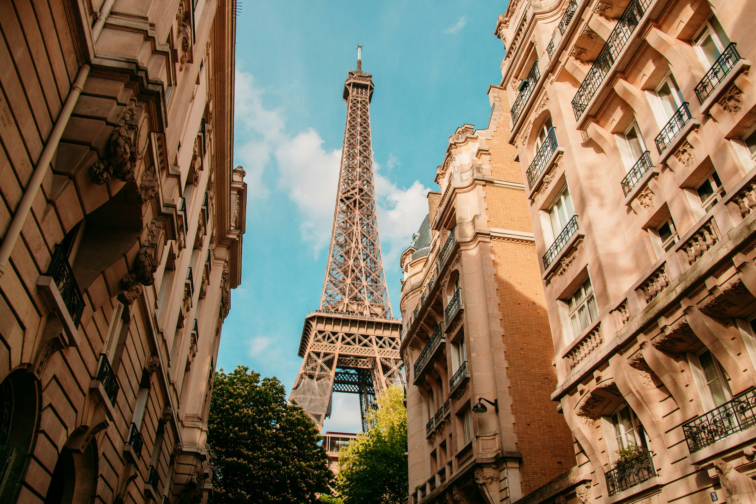 View of the Eiffel Tower between ornate Parisian residential buildings with balconies, under a partly cloudy blue sky.