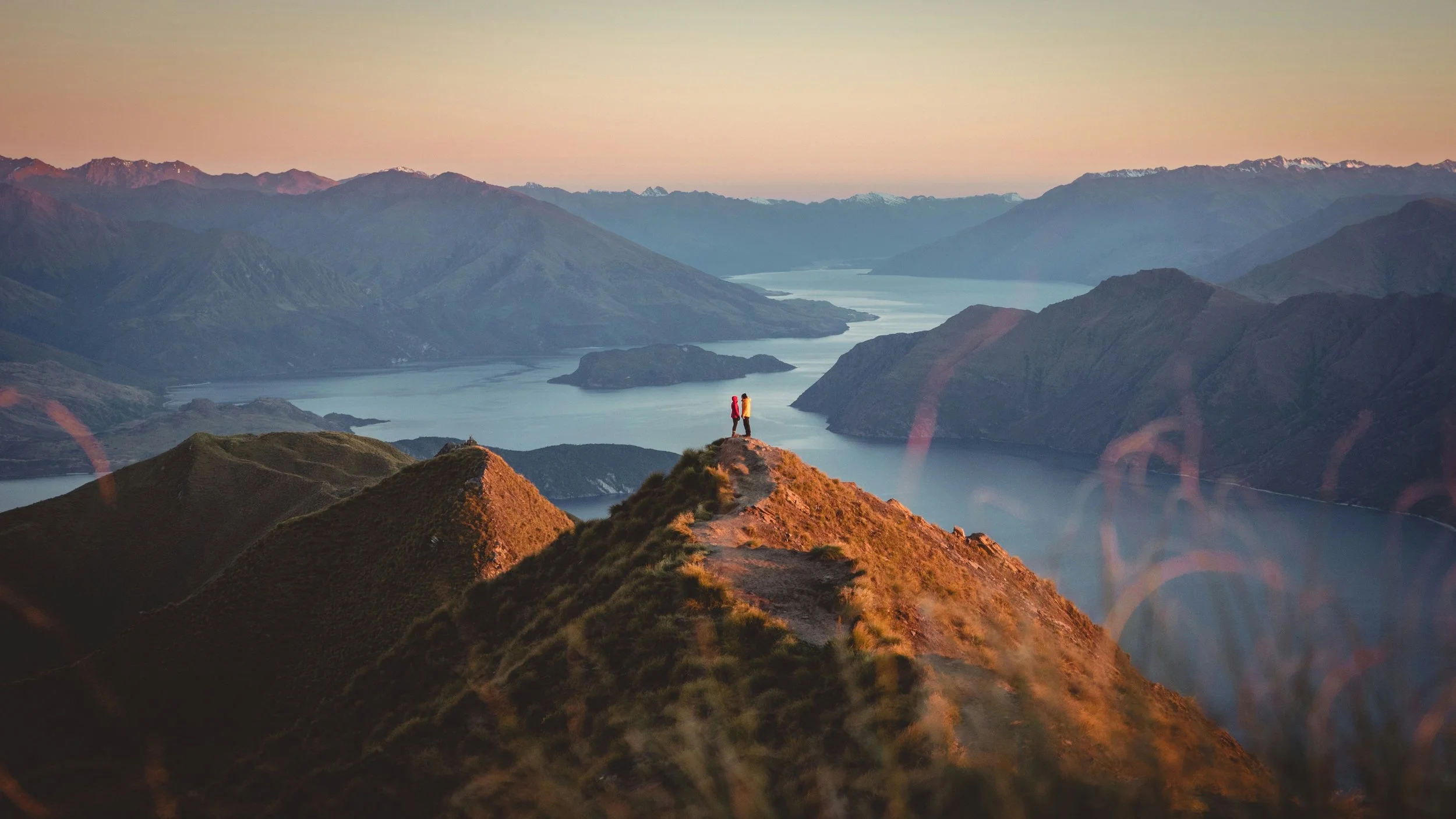 Two hikers standing on a mountain ridge overlooking a large lake surrounded by mountains at sunset.